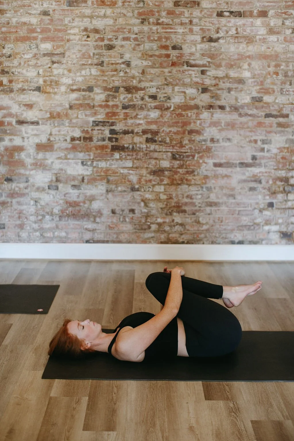 A young woman doing yoga on a mat against a brick wall, lying on their back with knees hugged to chest, wearing black athletic wear.