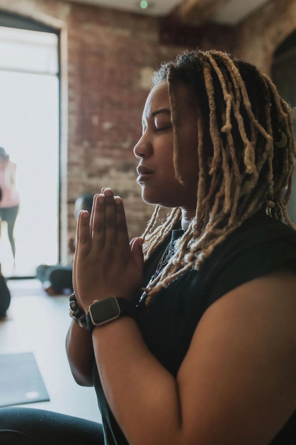 A woman sits in a yoga studio with prayer hands in front of her heart, eyes closed. On her wrist she wears an Apple Watch and beaded bracelets