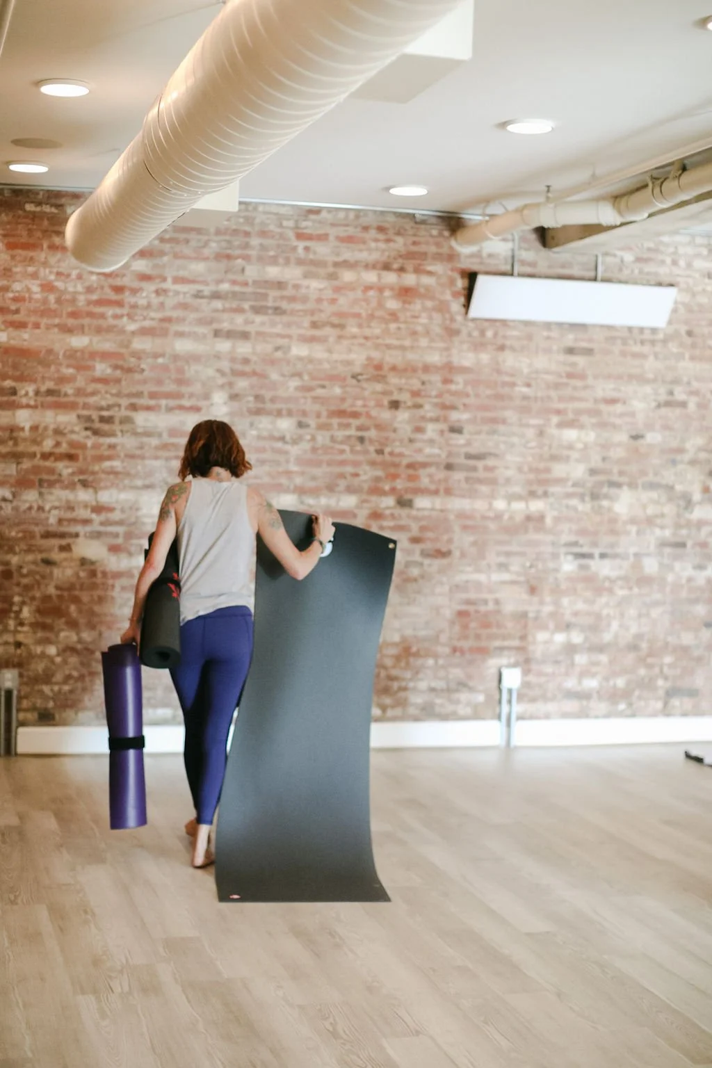 A woman holding a black yoga mat and rolled-up black yoga mat, looks for her place in a yoga studio with a brick wall background.
