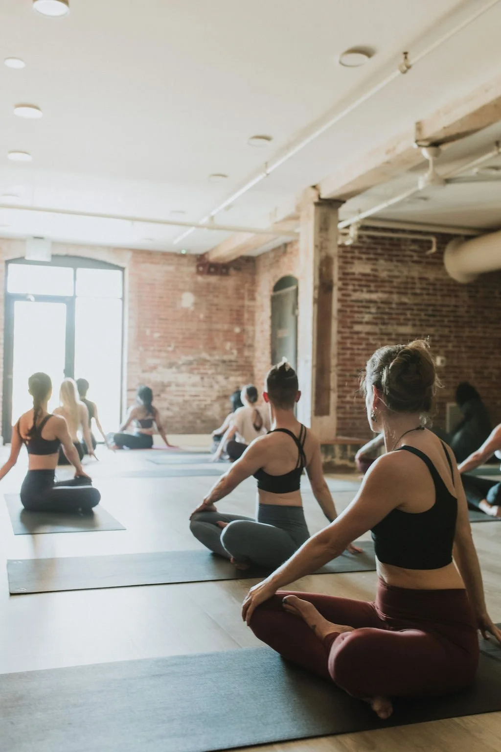 A yoga instructor leads a class with participants in a brick-walled studio. Participants are seated on yoga mats, twisting towards the light of the window