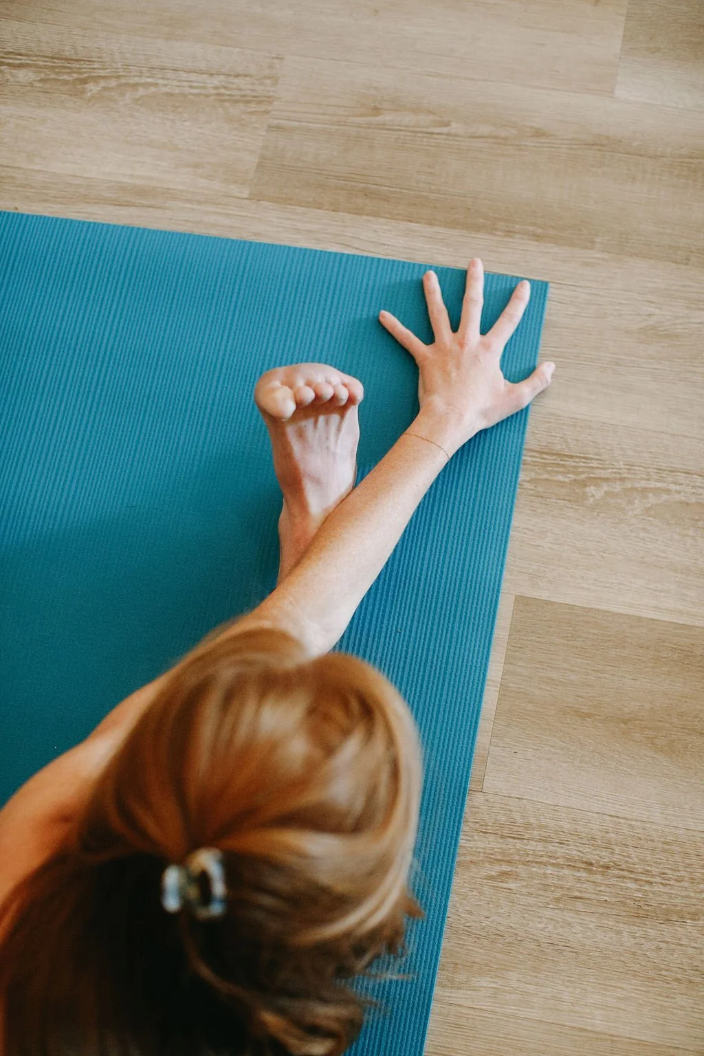 A red-haired woman stretching in janu sirsasana on a blue yoga mat on a wooden floor, with focus on extended arm and foot, viewed from above.