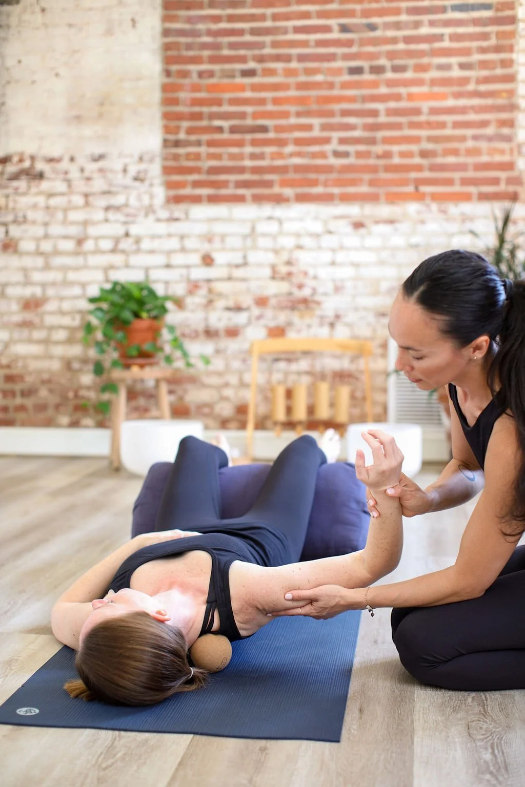 A woman lying on a yoga mat receiving physical therapy on her shoulder from a therapist.