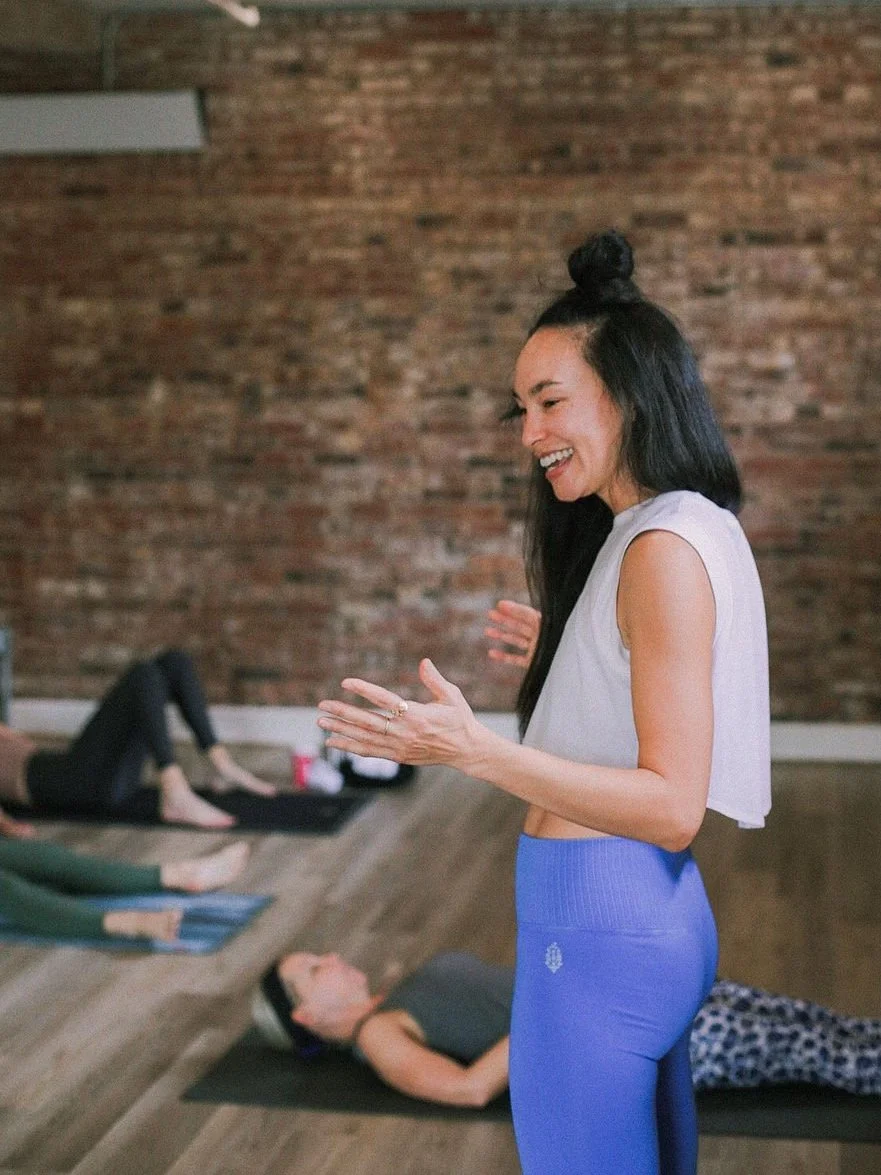 A woman leading a yoga class, smiling and gesturing with her hands, with beginner and experienced students lying on yoga mats in a studio with a brick wall background.