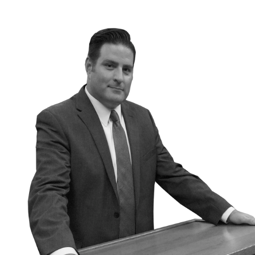 Black and white photo of a man in a suit and tie leaning on a table, looking at the camera.