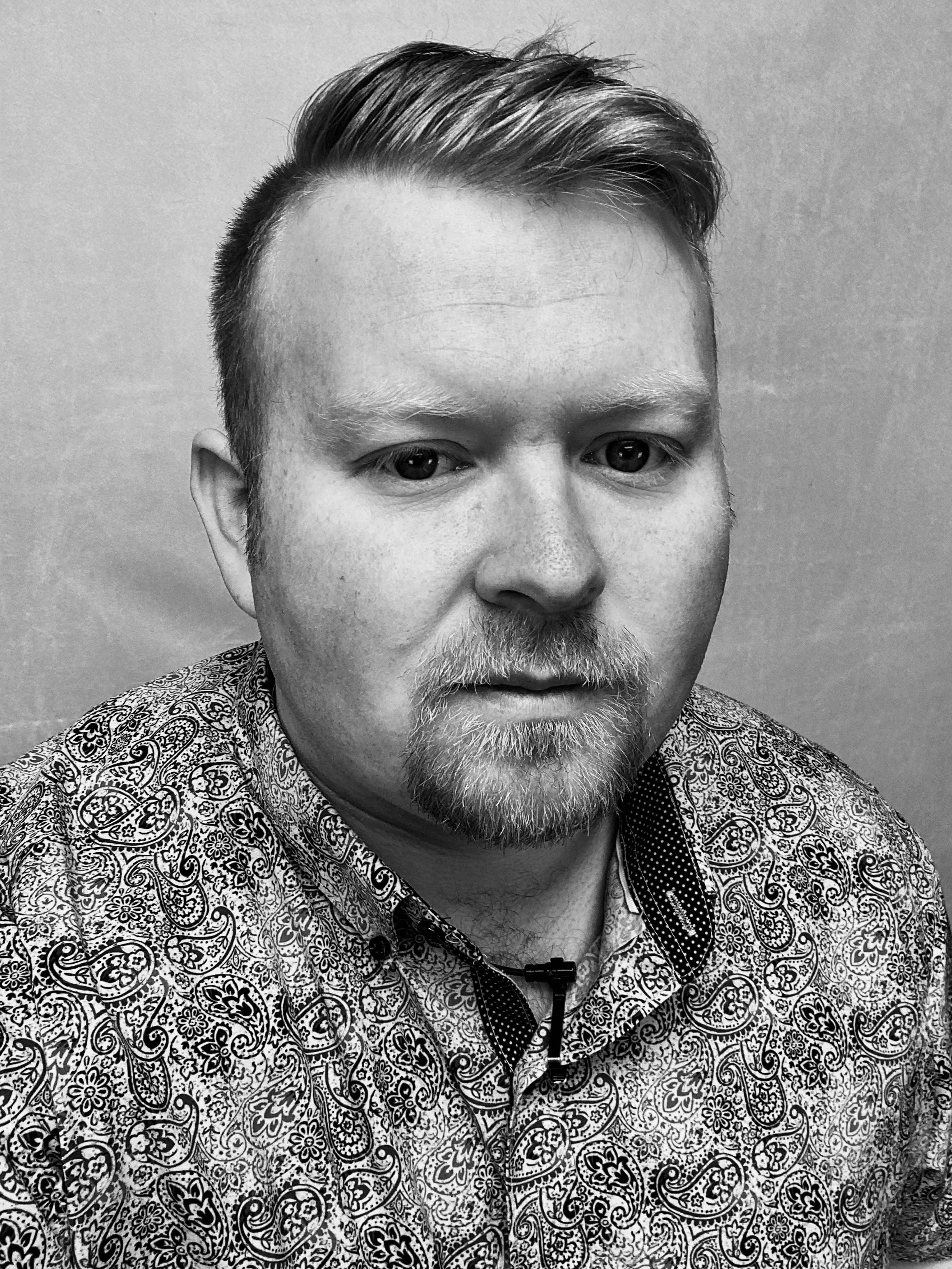 Black and white portrait of a young man with light-colored hair, facial hair, and wearing a patterned shirt.