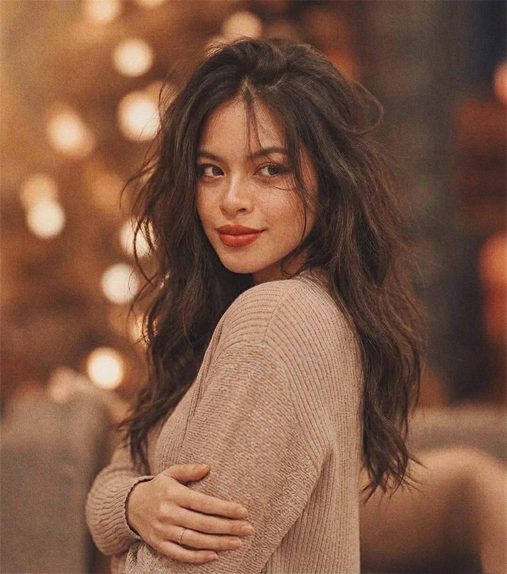 A woman with long, wavy dark hair looking over her shoulder, smiling slightly, wearing a beige sweater, with warm blurred lights in the background.