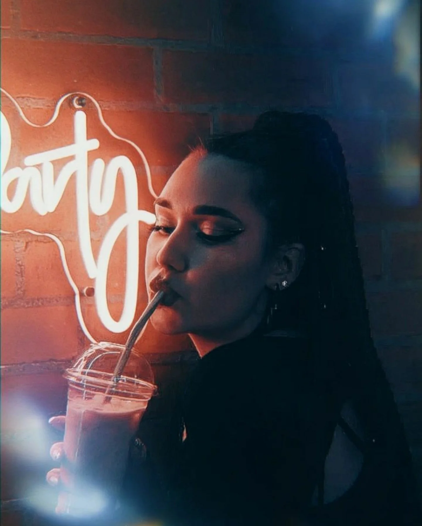 A woman with dark hair styled in an updo, wearing earrings, is drinking from a straw in a glass with a dark beverage, in front of a neon sign that says 'eat'. The background is a brick wall.