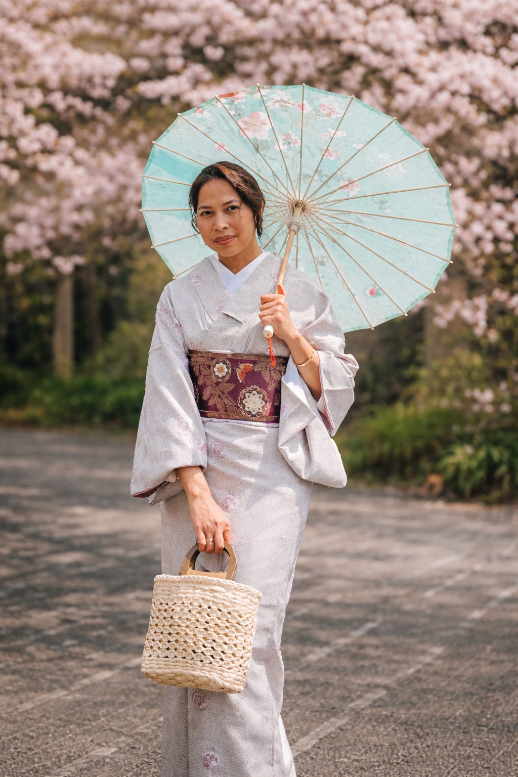 A woman wearing a traditional Japanese kimono holding a parasol and a woven bag, standing outdoors among cherry blossom trees.