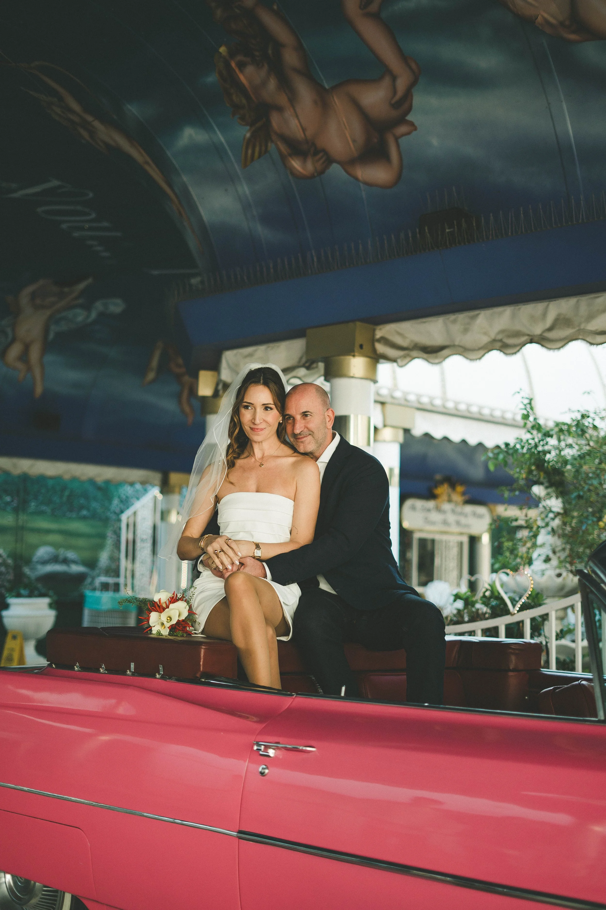 A bride and groom sitting on the back of a pink vintage convertible car in front of a carousel with painted cherubs and clouds, during a wedding celebration.