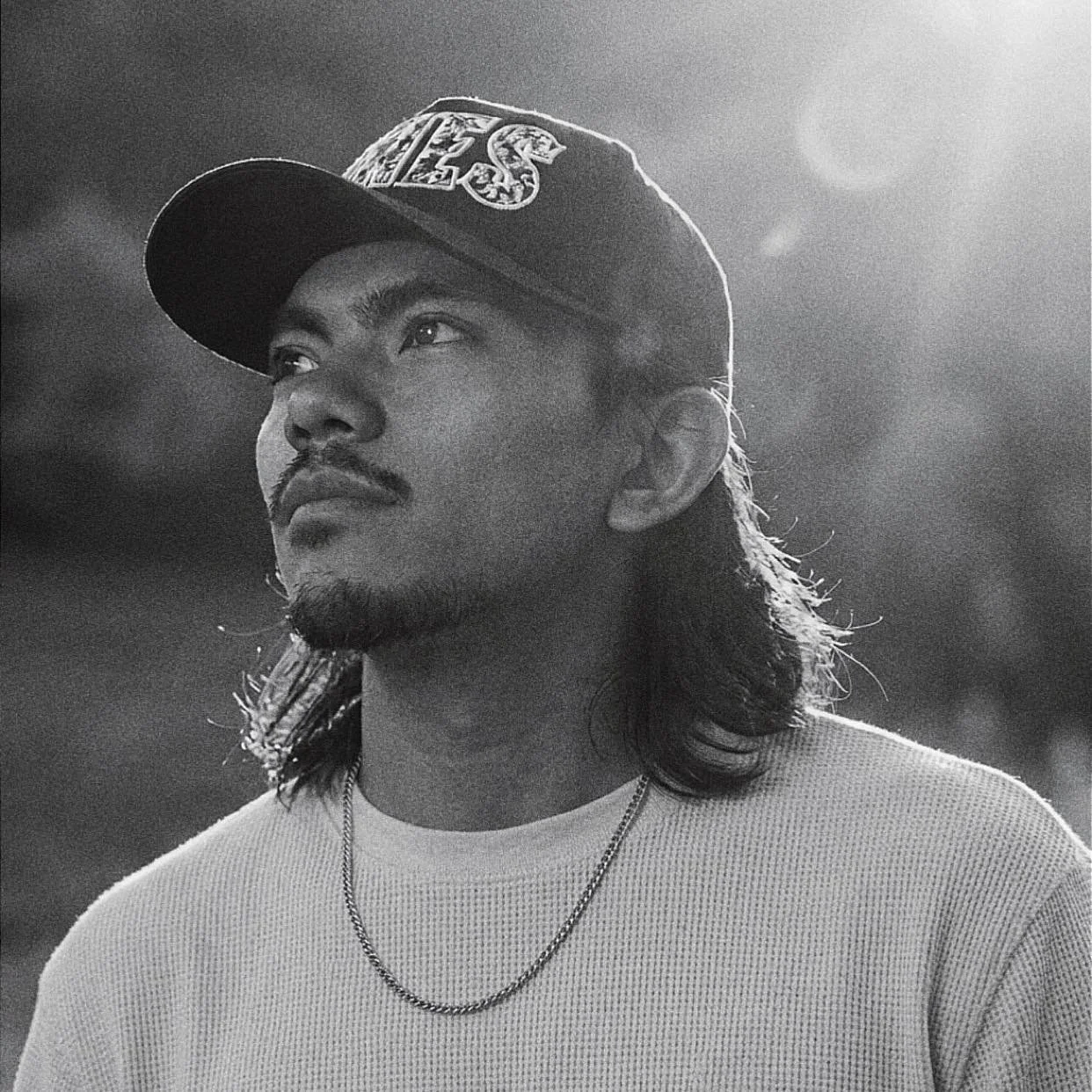 Black and white portrait of a young man with long hair, a mustache, and a goatee, wearing a cap with a logo, a light-colored sweater, and a chain necklace, looking pensively to the side.