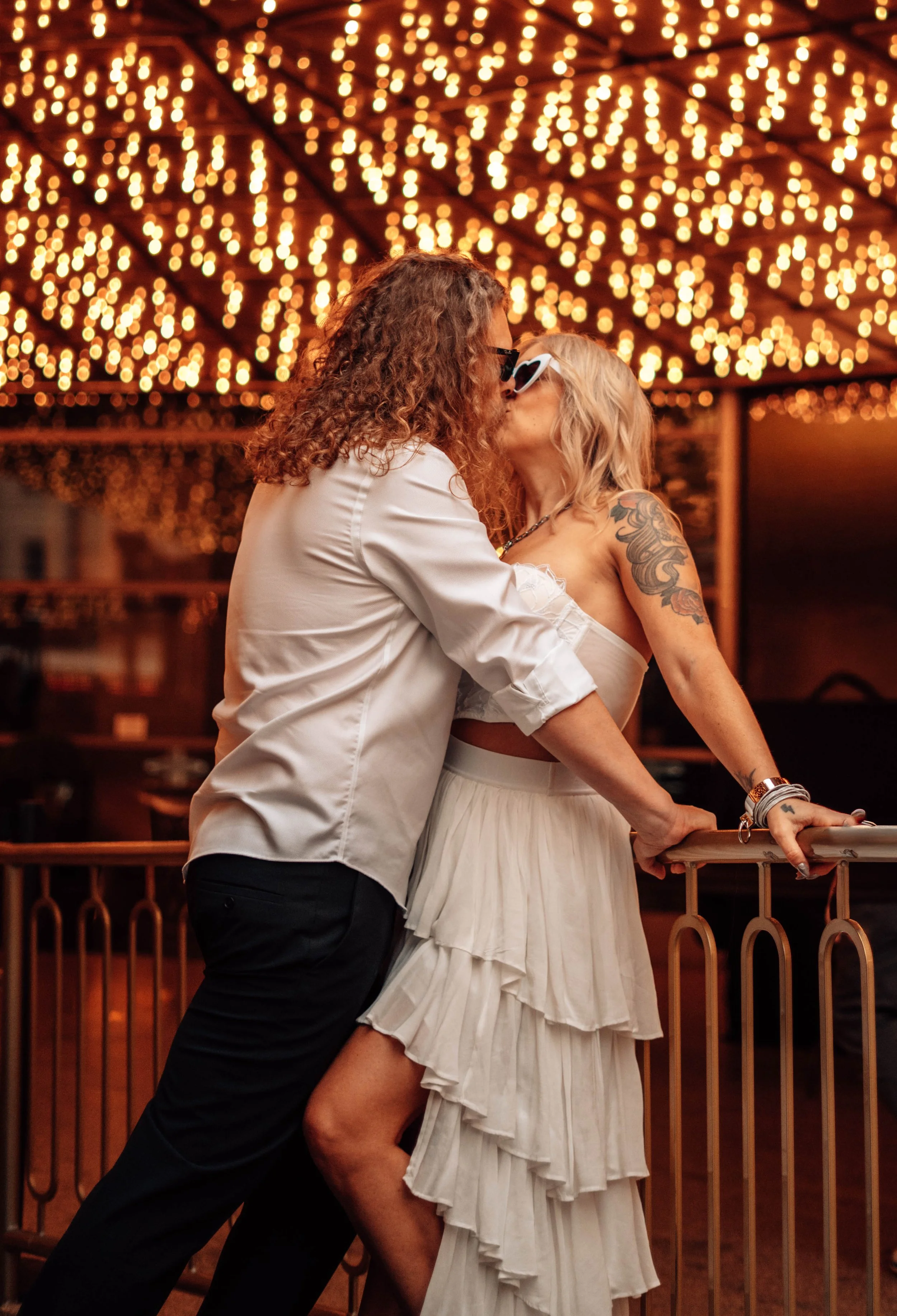 A couple kissing under string lights at night on a balcony with a wooden railing.