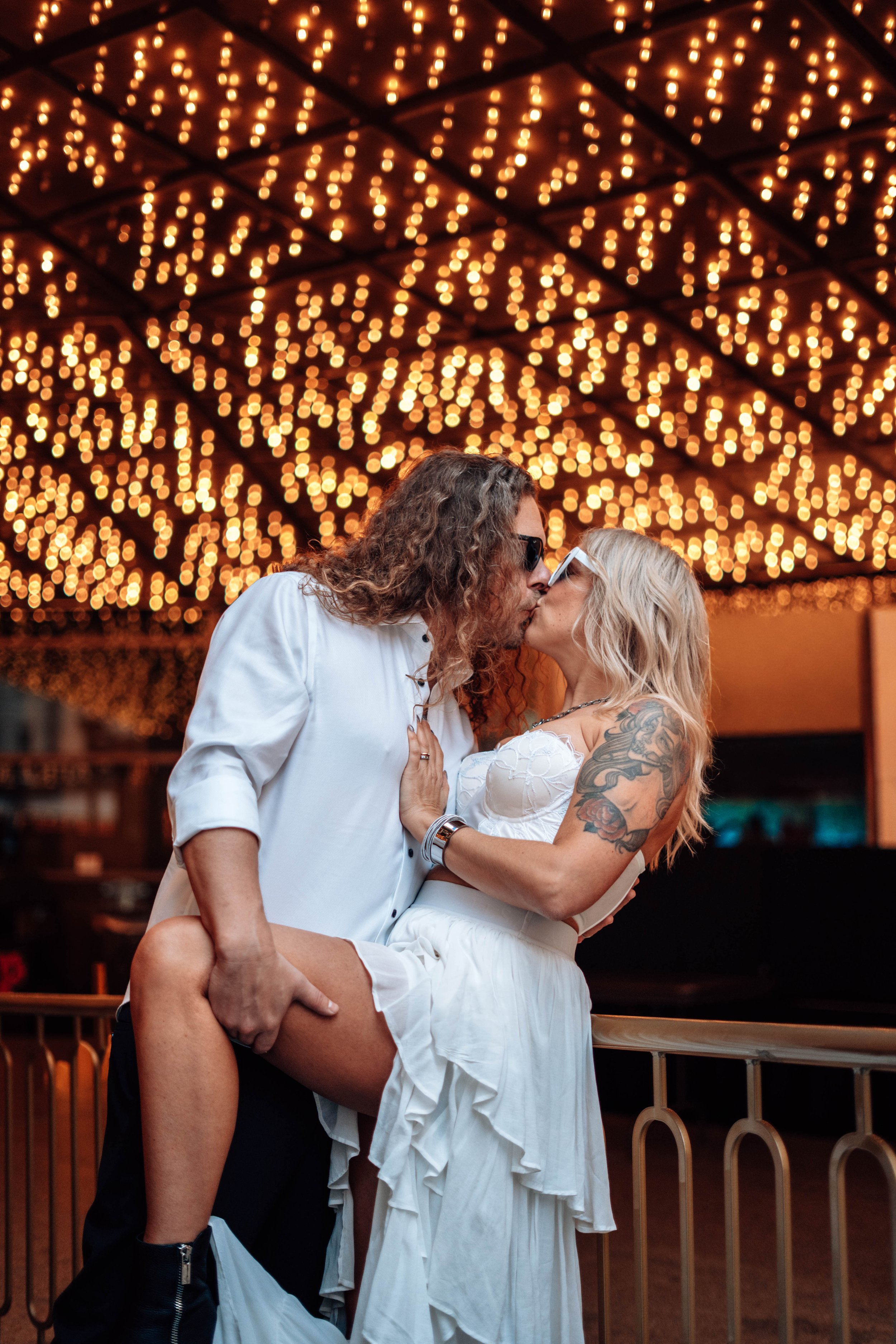 A couple kissing in a romantic moment with string lights hanging overhead in a dimly lit setting.