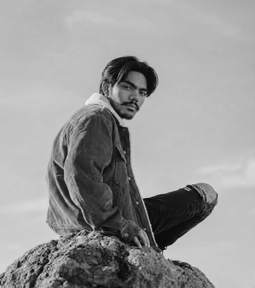 A man with dark hair and a beard, wearing a denim jacket and ripped jeans, sitting on a rock outdoors, looking at the camera in black and white.