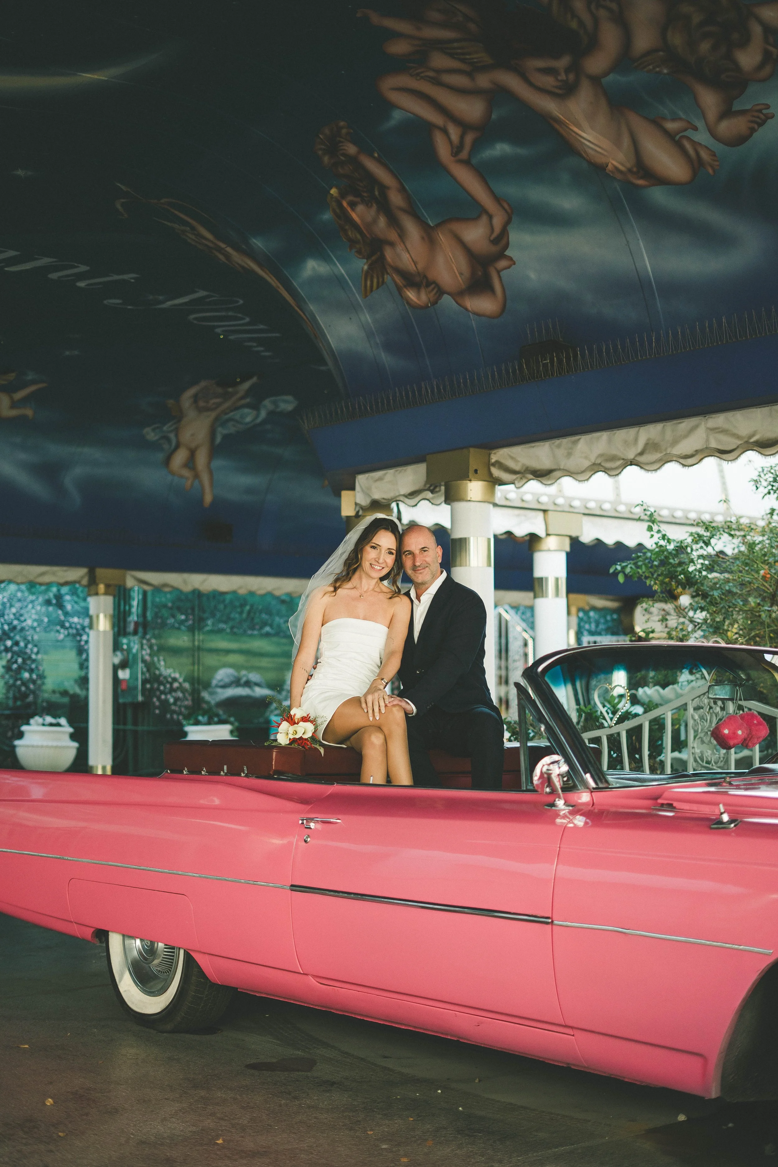 A bride and groom sitting in a pink vintage convertible car, inside an amusement park or fairground, with a painted ceiling featuring cherubs.