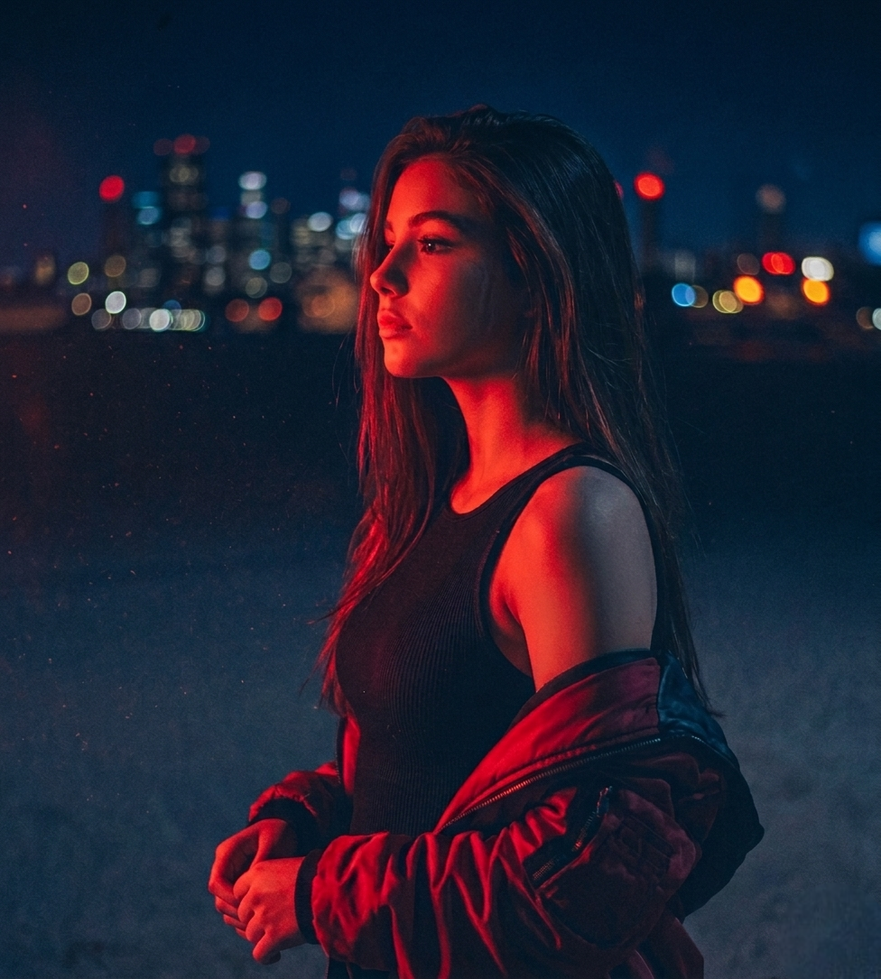 A woman with long dark hair stands outdoors at night against a city skyline with blurred lights, wearing a black sleeveless top and a red jacket off her shoulders, illuminated by red lighting.