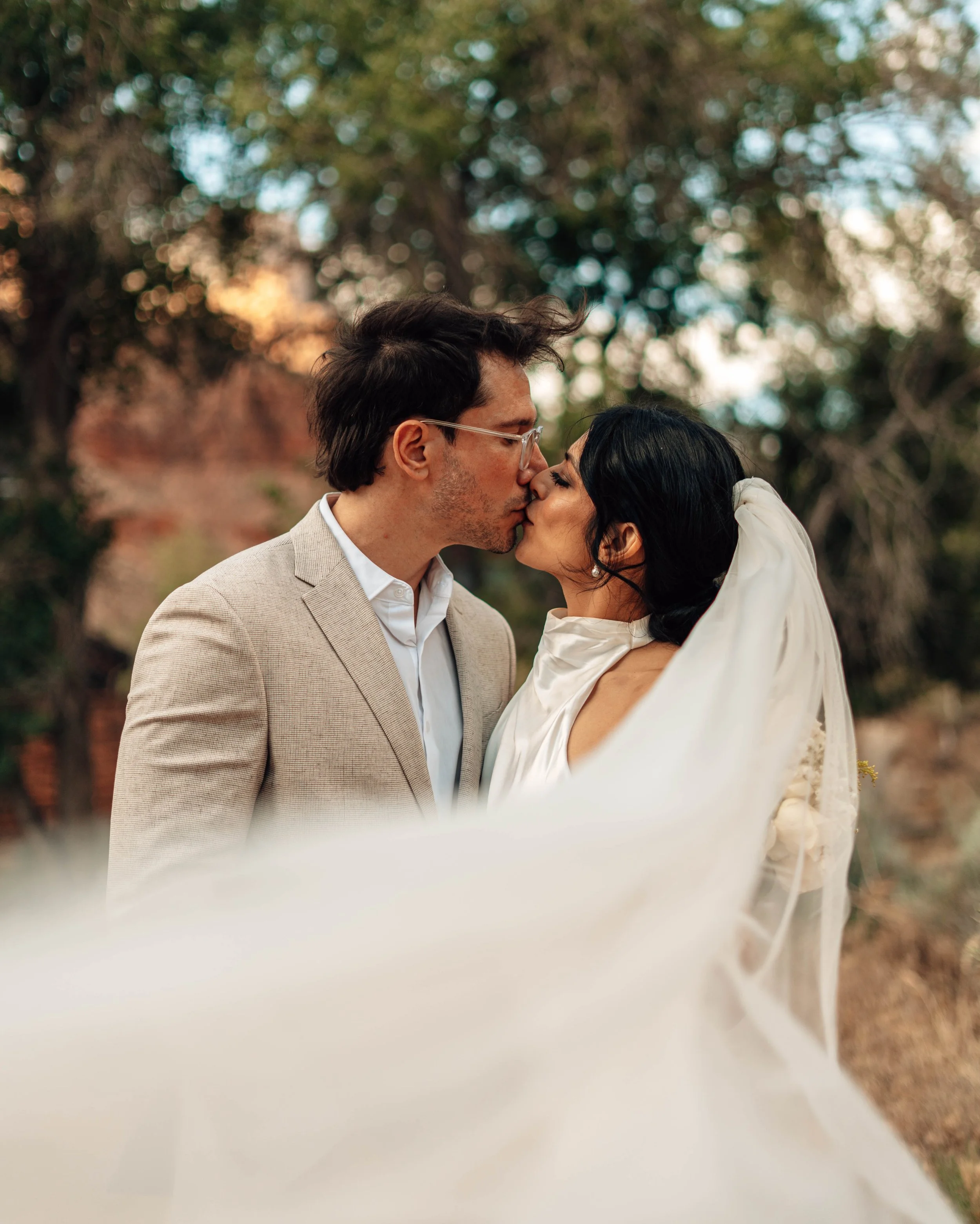 A couple sharing a kiss outdoors with blurred trees and sky in the background, partially obscured by a white cloth in the foreground.