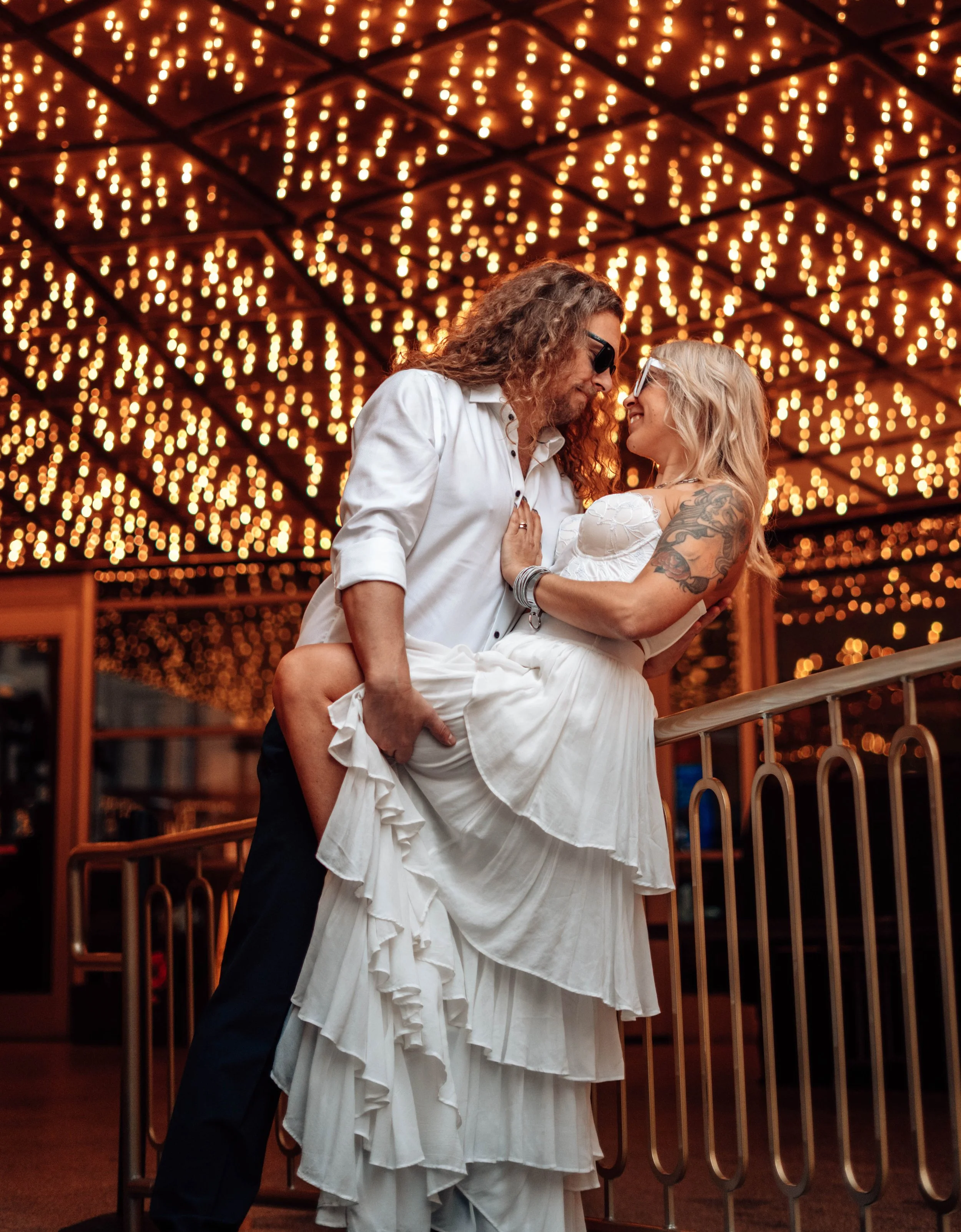A couple dancing closely under ceiling with string lights, with man wearing sunglasses and woman with tattoos on her arm, in a warm, festive setting.
