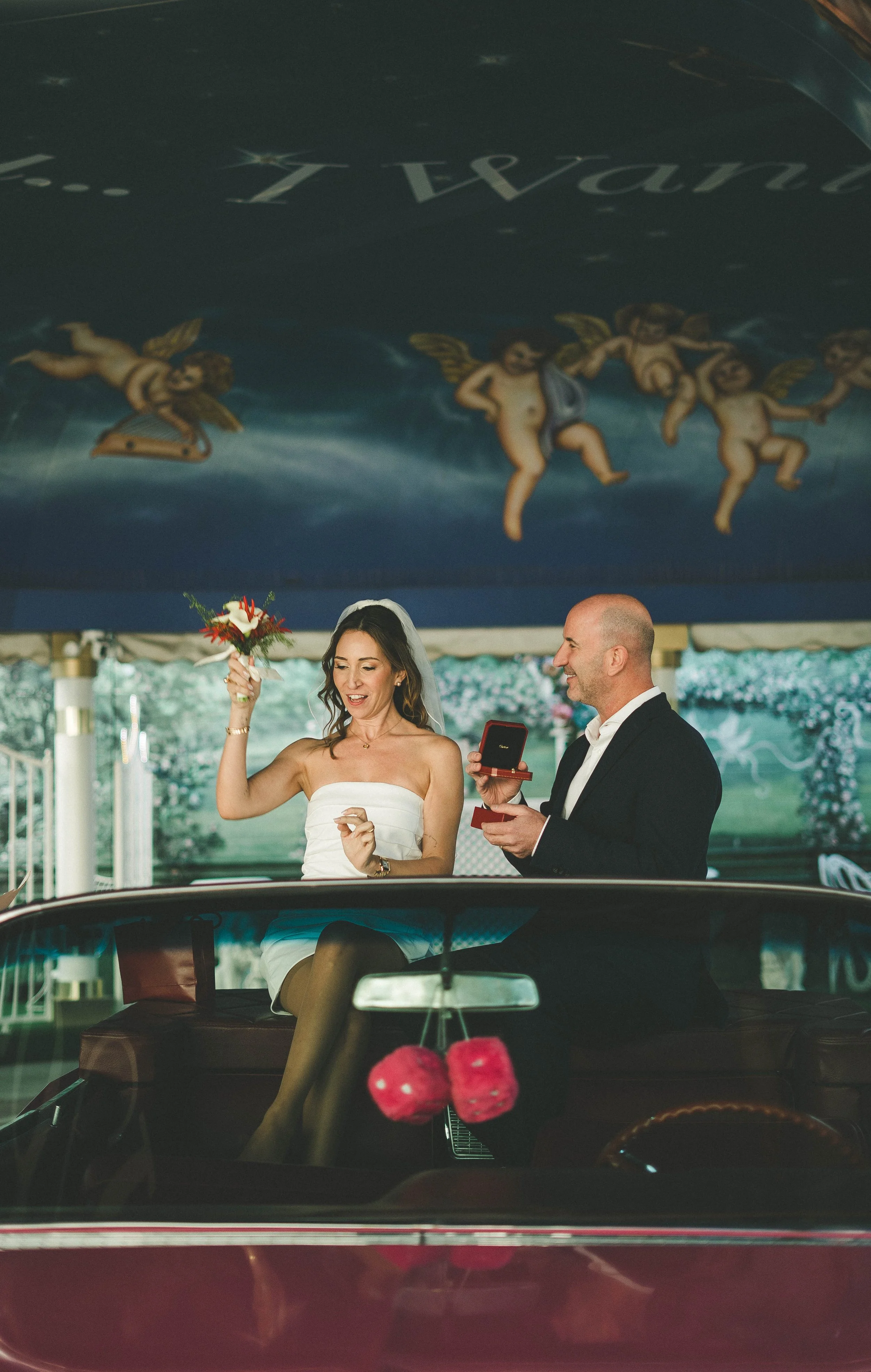 A bride and groom are standing inside a car during a wedding ceremony. The bride is holding a bouquet of flowers and smiling, while the groom is holding a ring box and smiling at her. The photo is taken from outside the car, with a reflection on the 