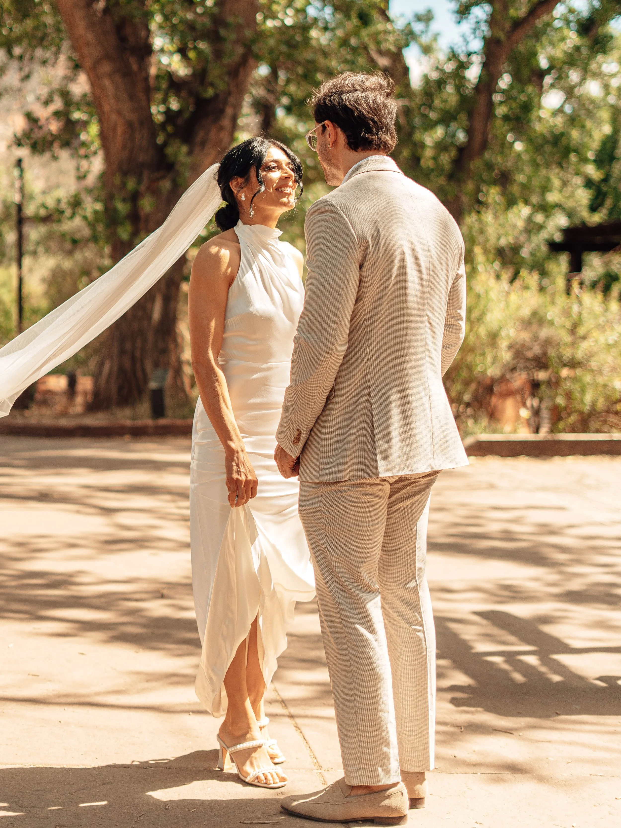 A bride and groom stand outdoors under a large tree, smiling at each other, holding hands during a wedding ceremony.