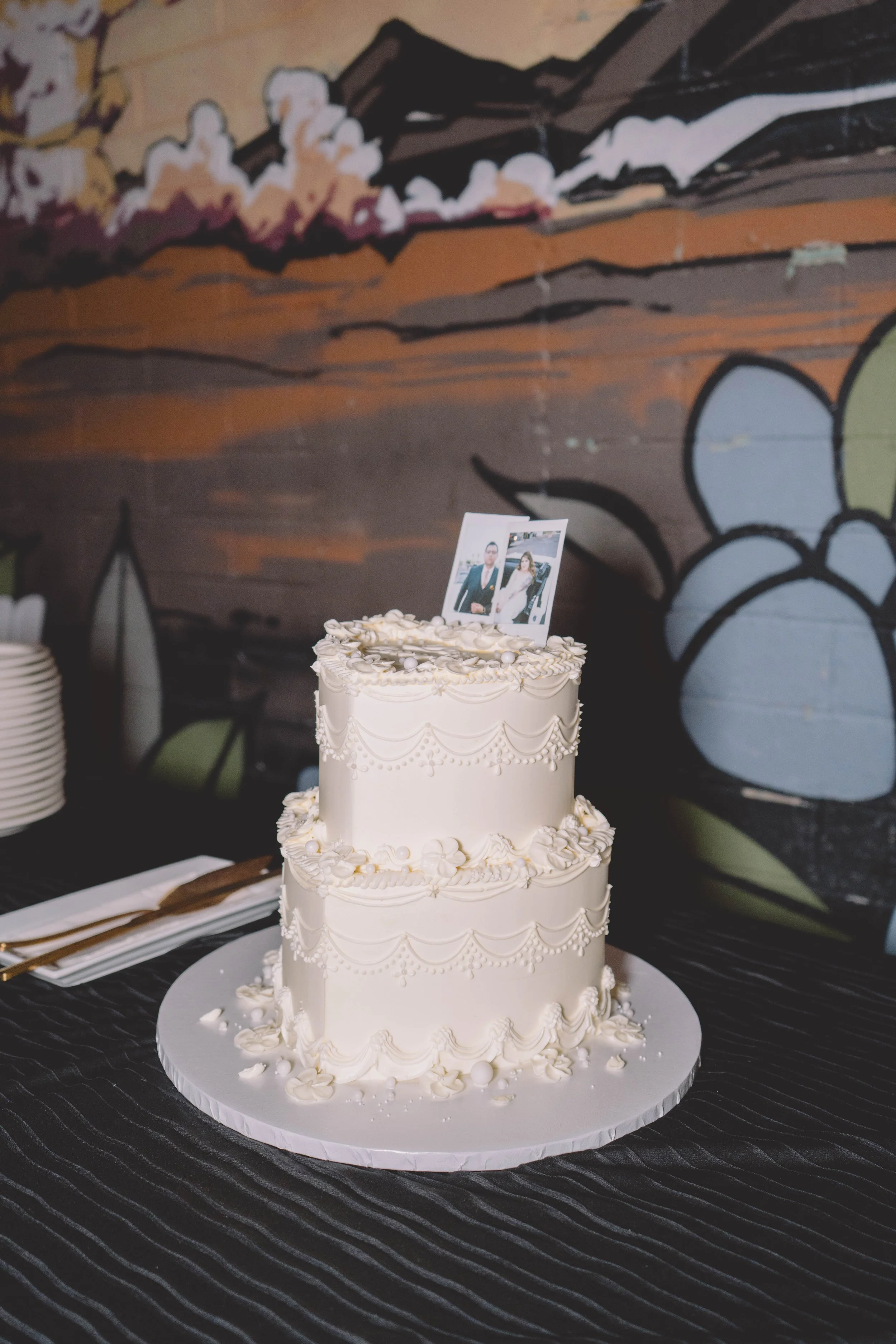 Three-tiered white wedding cake decorated with piped icing and small white edible pearls, topped with a photograph of a bride and groom, on a white cake board on a black tablecloth with a striped pattern, against a background of graffiti art on a brick wall.