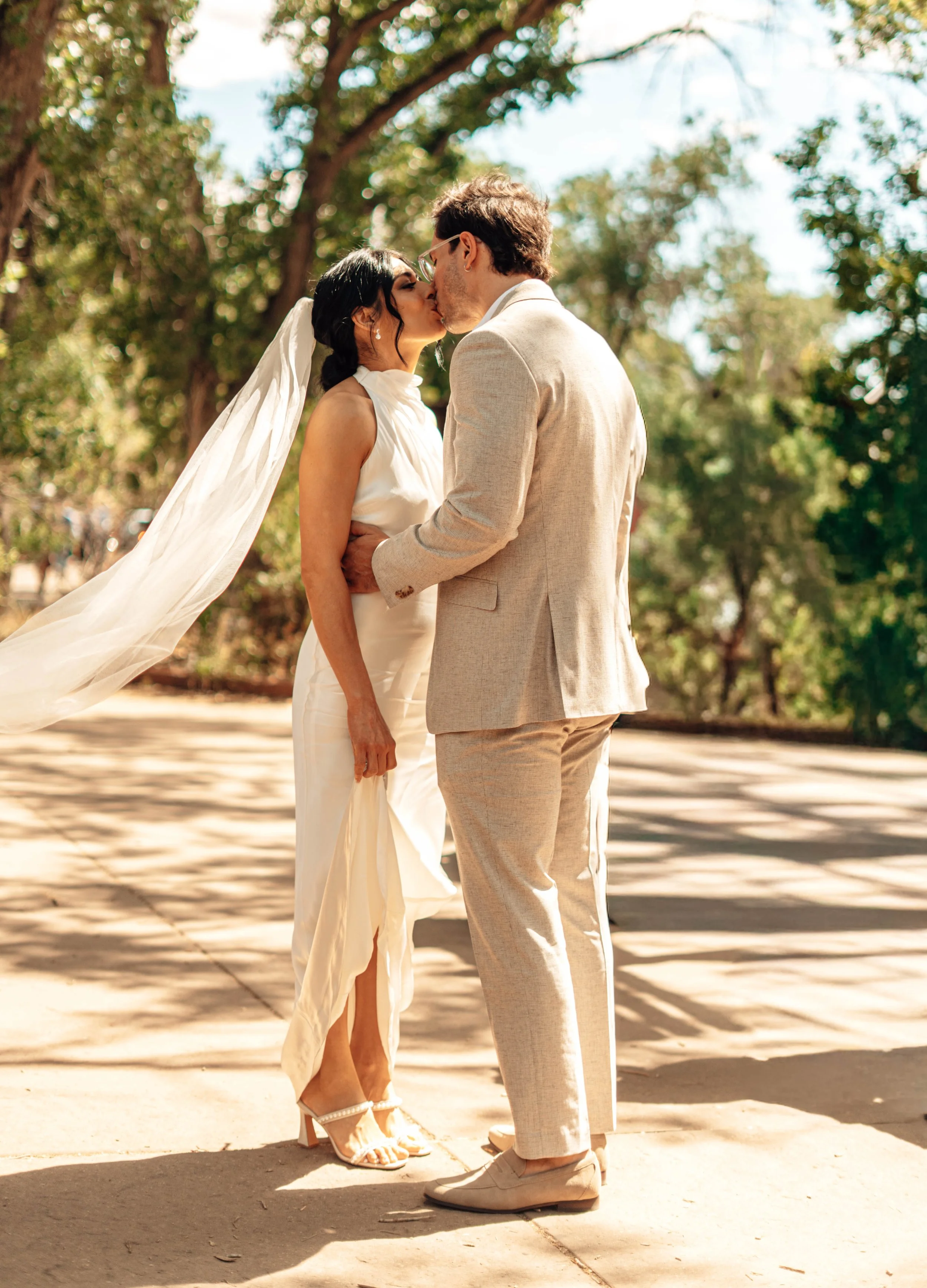 A couple is kissing outdoors during daytime. The woman is wearing a white dress and heels, with a veil flowing behind her. The man is dressed in a light-colored suit with matching pants and shoes. They are standing on a sidewalk with trees in the bac