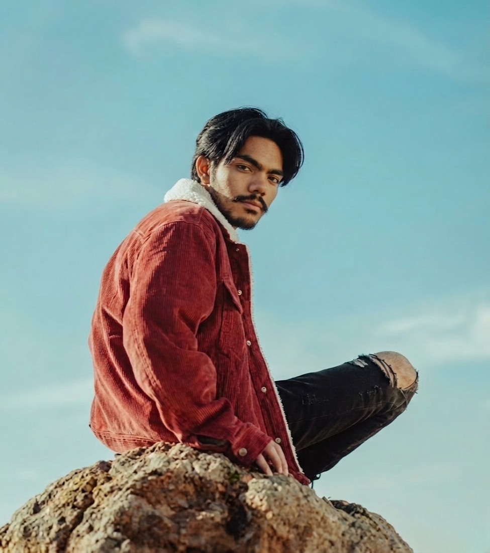 A man with black hair and a beard sitting on a large rock outdoors, wearing a red jacket with a white or light-colored fleece collar, and ripped black jeans, under a clear blue sky.