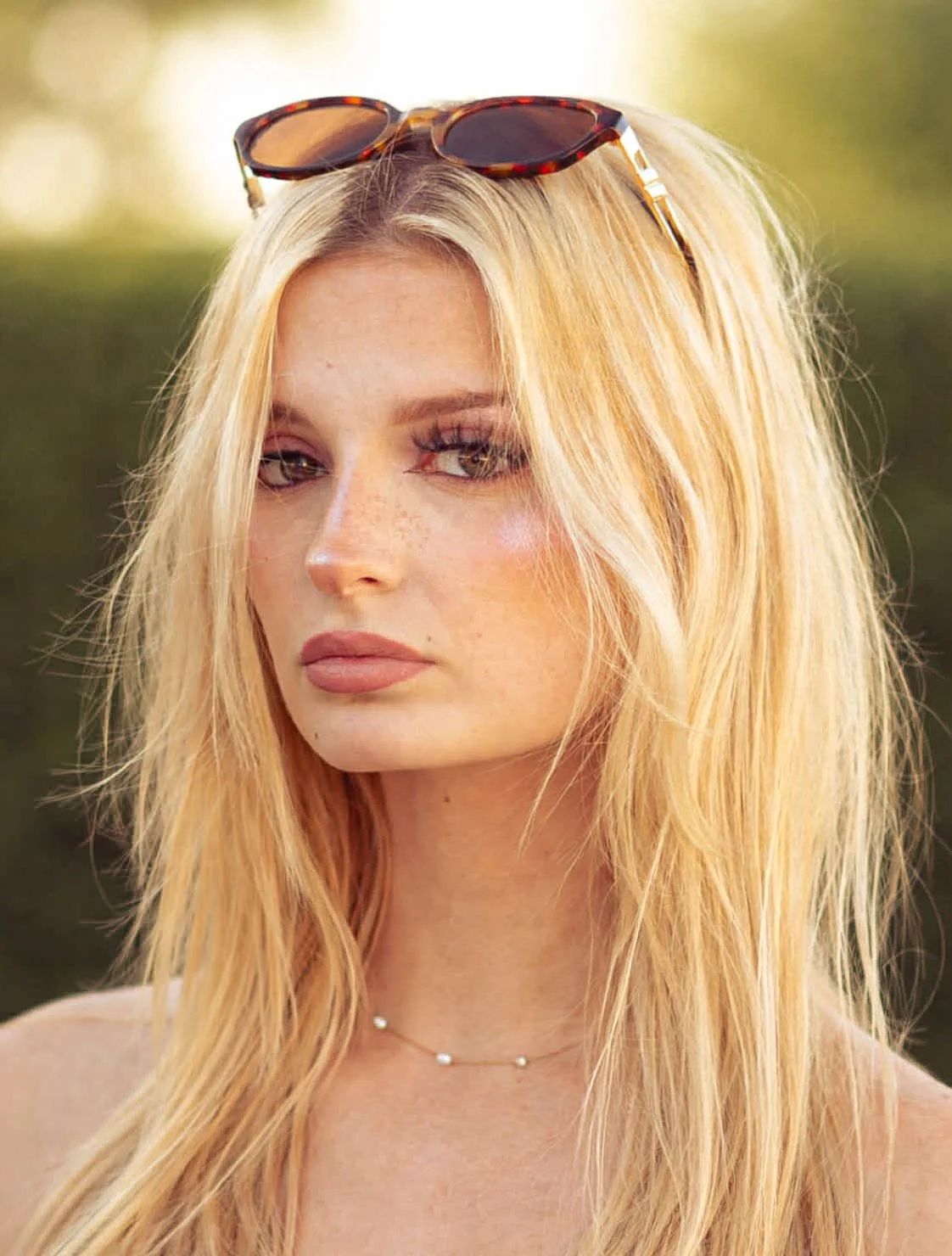 Close-up of a young woman with long blonde hair, wearing sunglasses on her head, a delicate necklace, and a neutral expression.