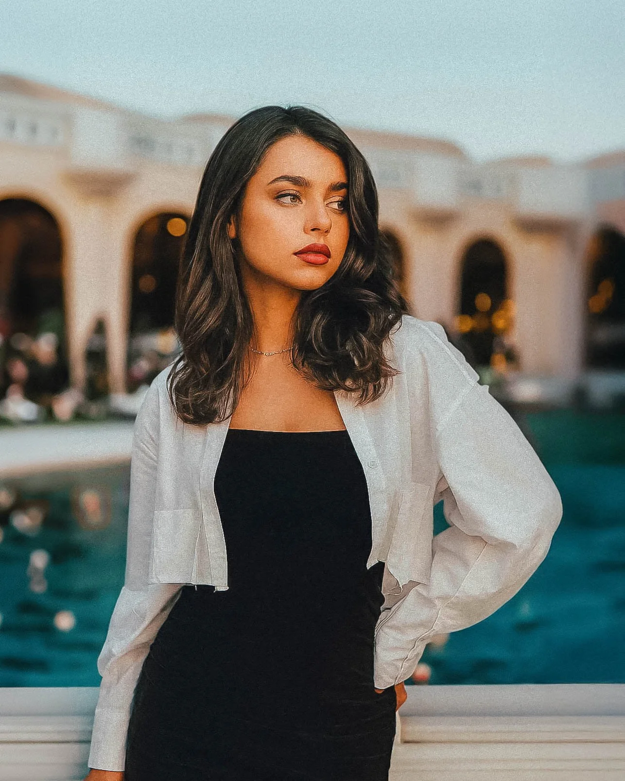 A young woman with dark wavy hair wearing a black dress and white shirt standing outdoors near water with a building in the background during sunset.