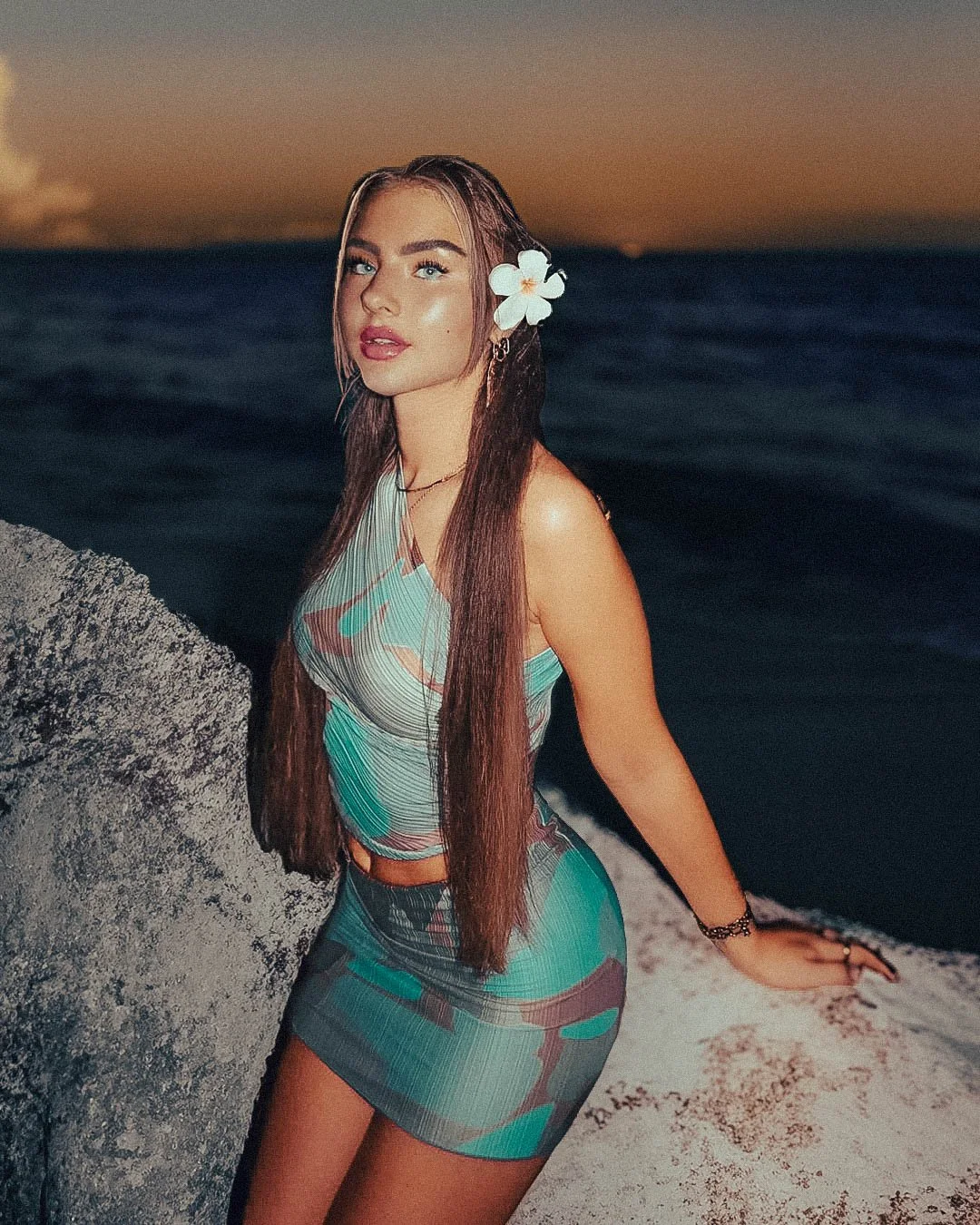 A young woman standing on a beach at sunset, wearing a colorful, patterned dress and a white flower in her hair.