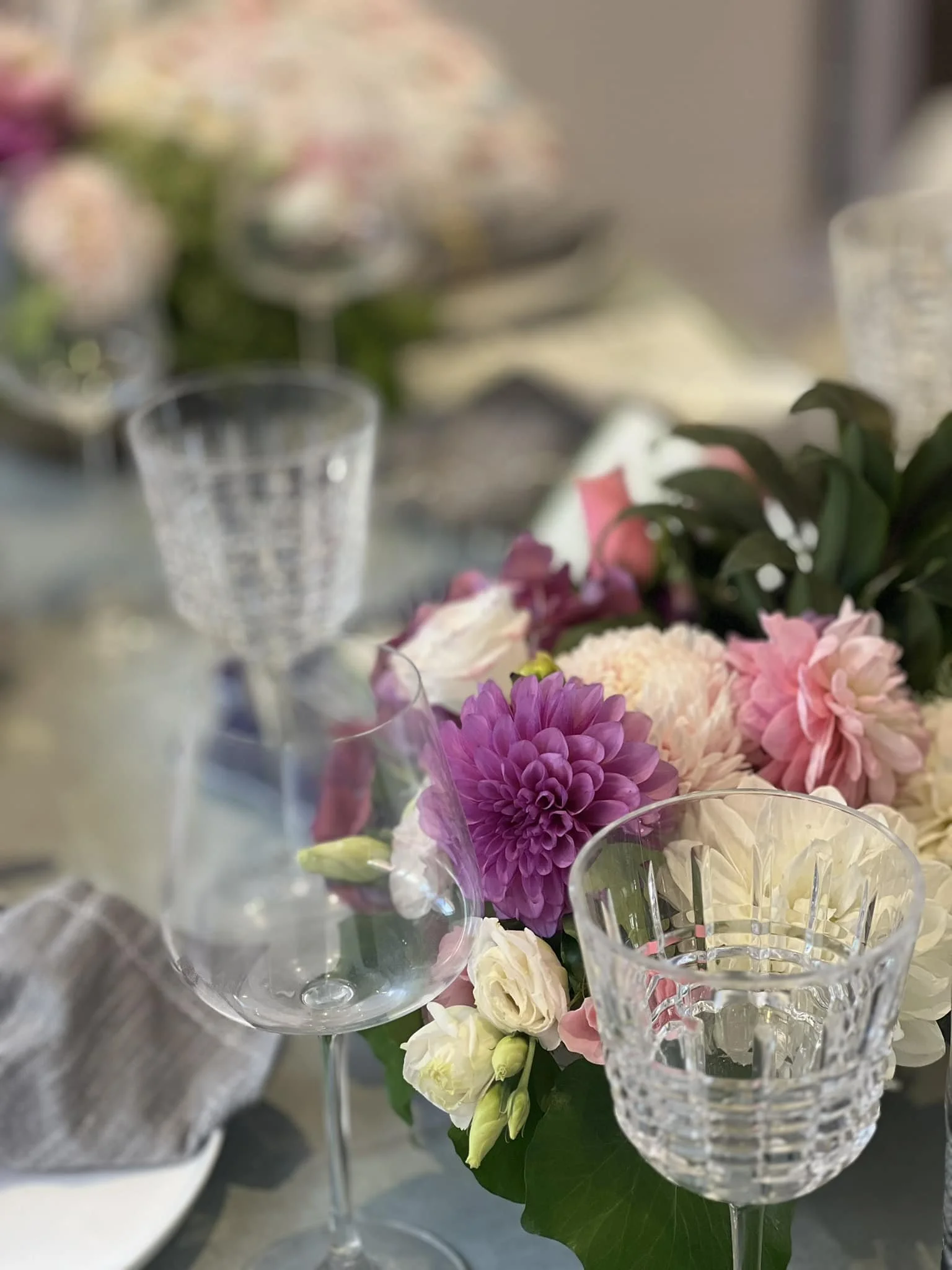 A floral centerpiece with pink, purple, and white flowers on a table, with crystal and glassware around it.