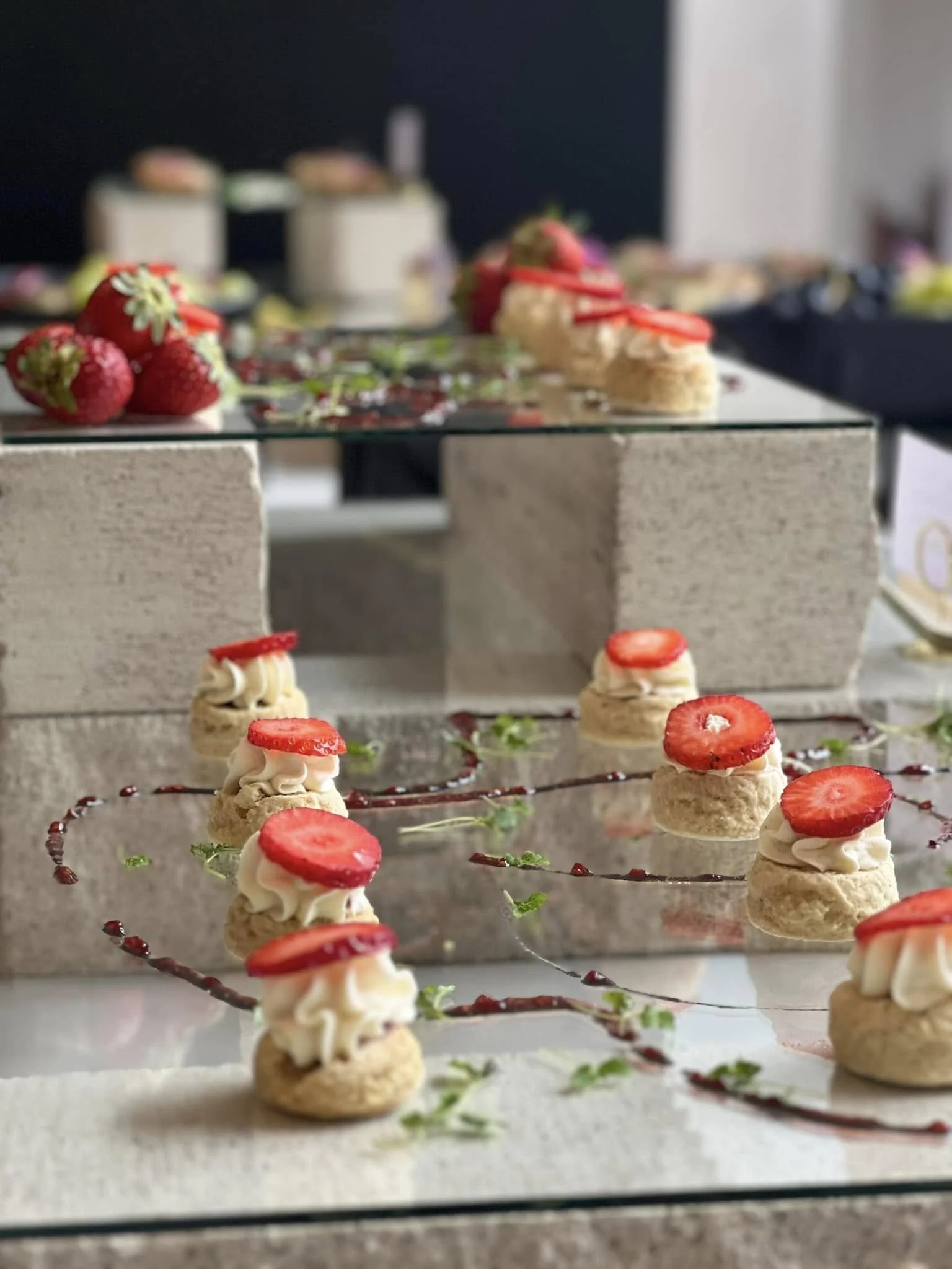 Assorted desserts including strawberry cream puffs topped with strawberry slices, strawberries, and cake on a glass display table with a decorative garnish of strawberry sauce and mint leaves.