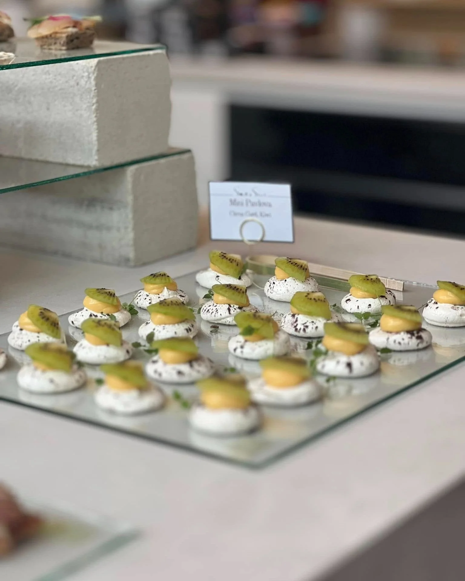 Mini pavlova dessert topped with sliced kiwi and a small dollop of yellow cream, arranged on a glass display tray at a bakery or cafe.