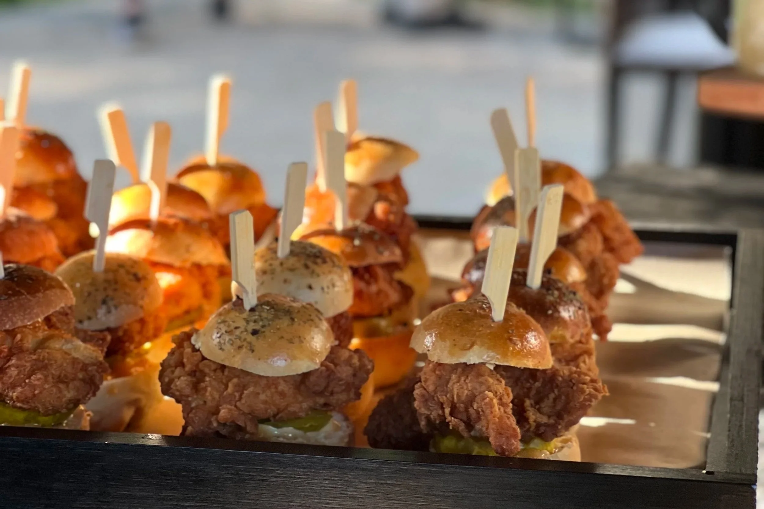 Close-up of several mini sliders with fried chicken, served on a wooden tray with toothpicks for a canape pass around.