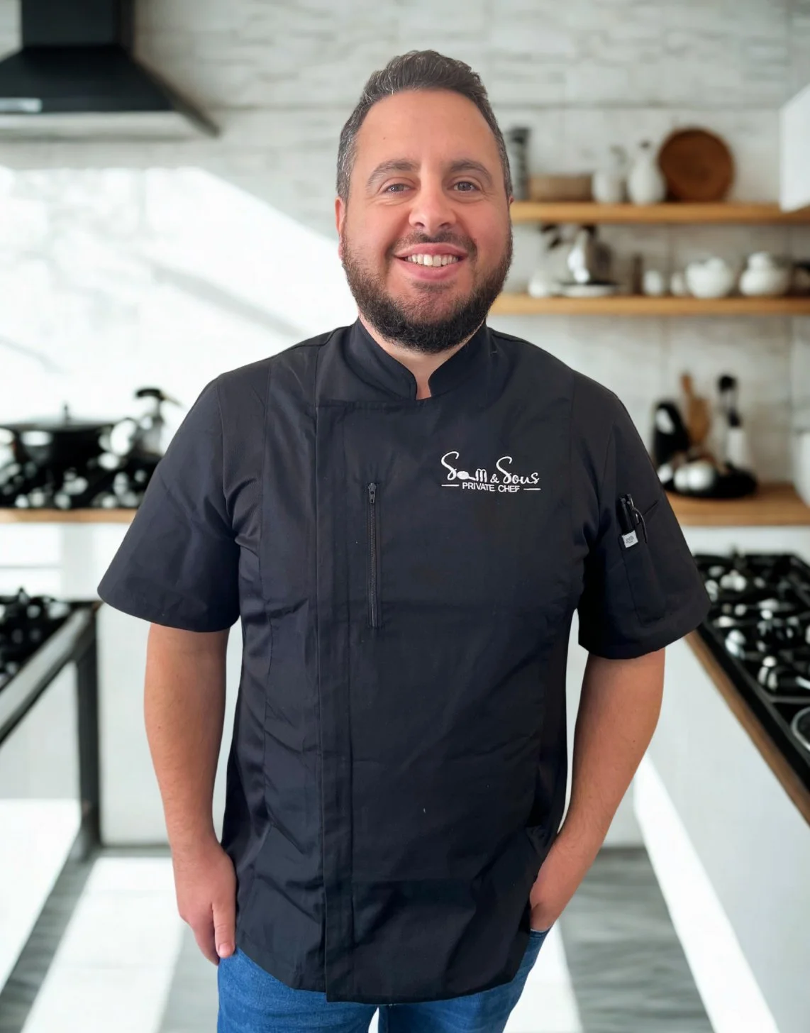 Man in a black chef's coat smiling in a modern kitchen.