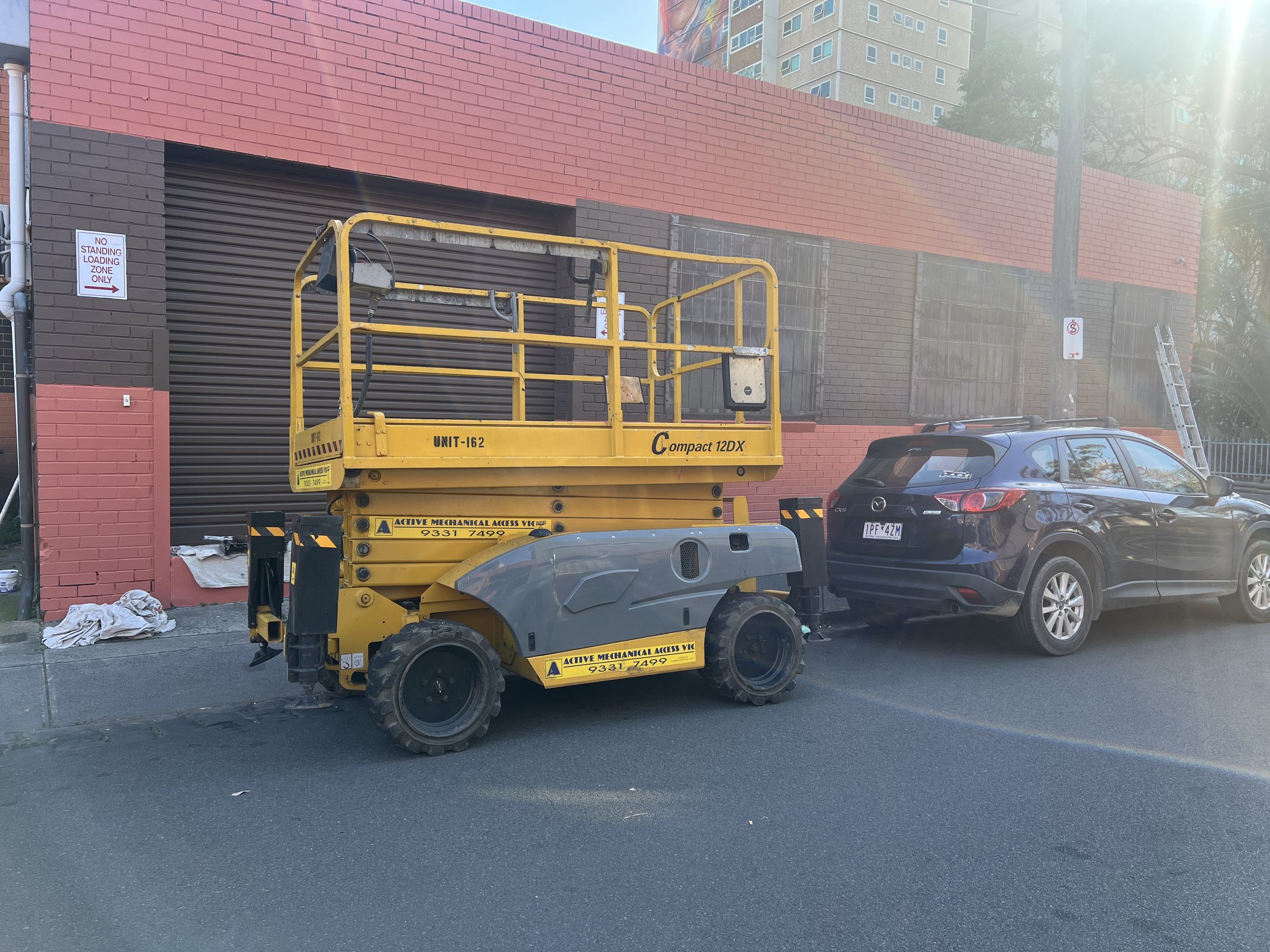 A yellow scissor lift parked on the street beside a car near a brick building.