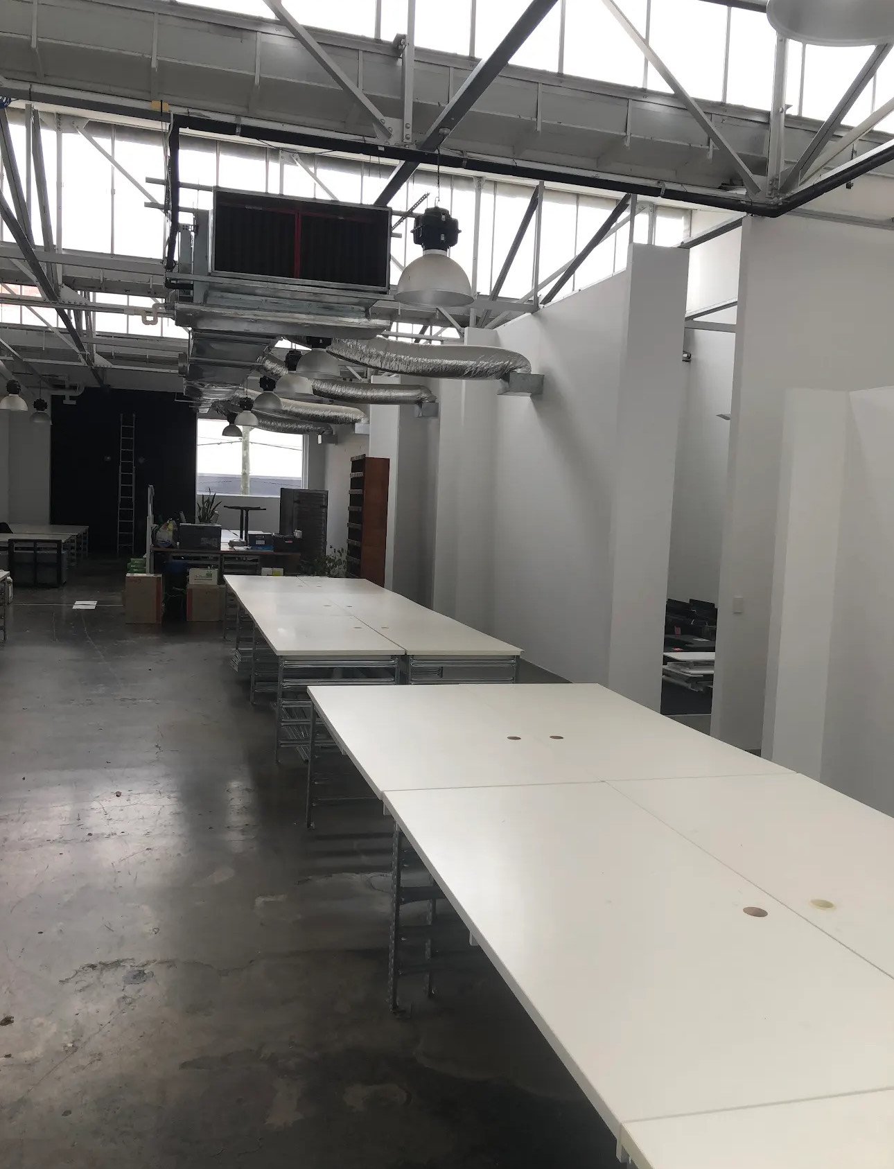 Industrial office space with white desks, exposed ceiling beams, ductwork, and large windows letting in natural light.