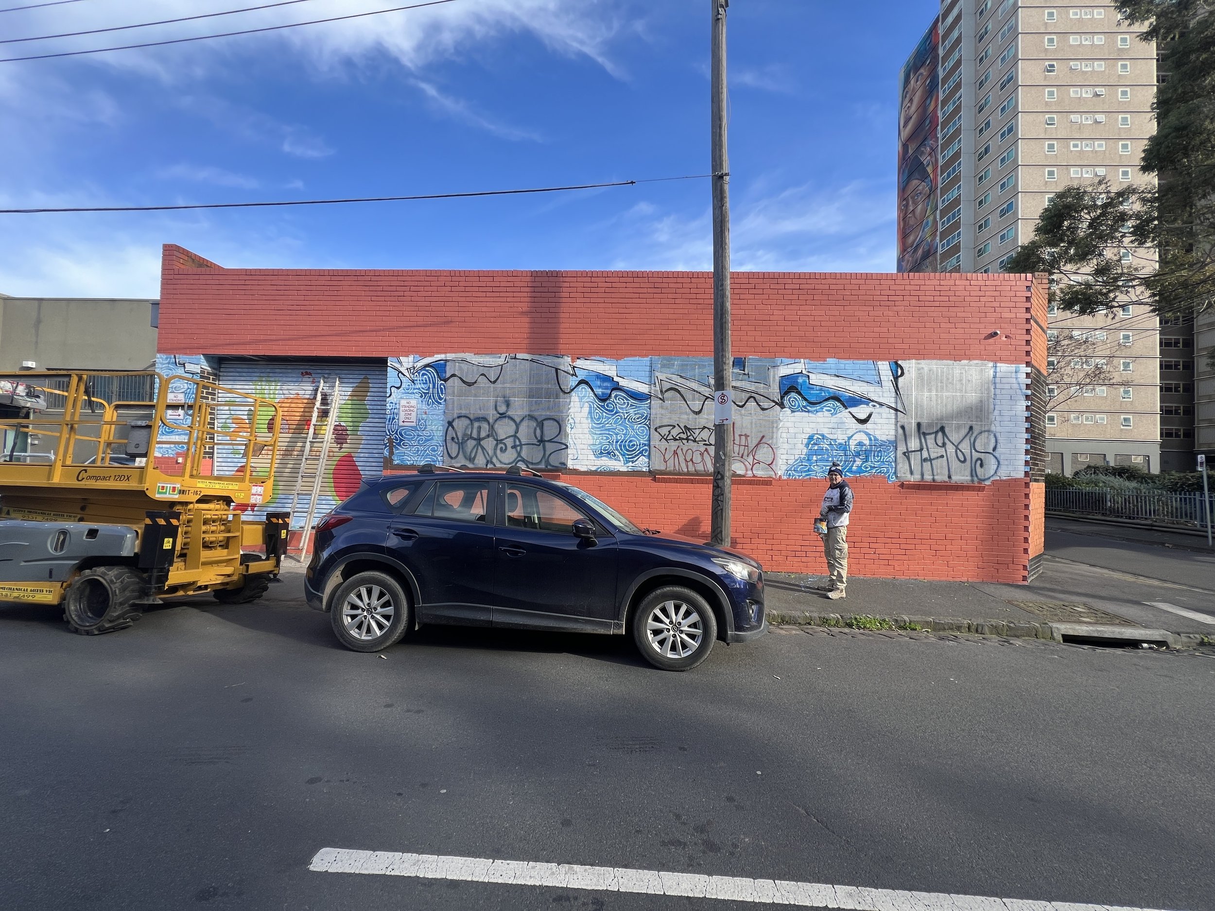Red brick building with graffiti on metal panels, parked black car, yellow lift, person standing on sidewalk, tall apartment building in the background, clear blue sky.