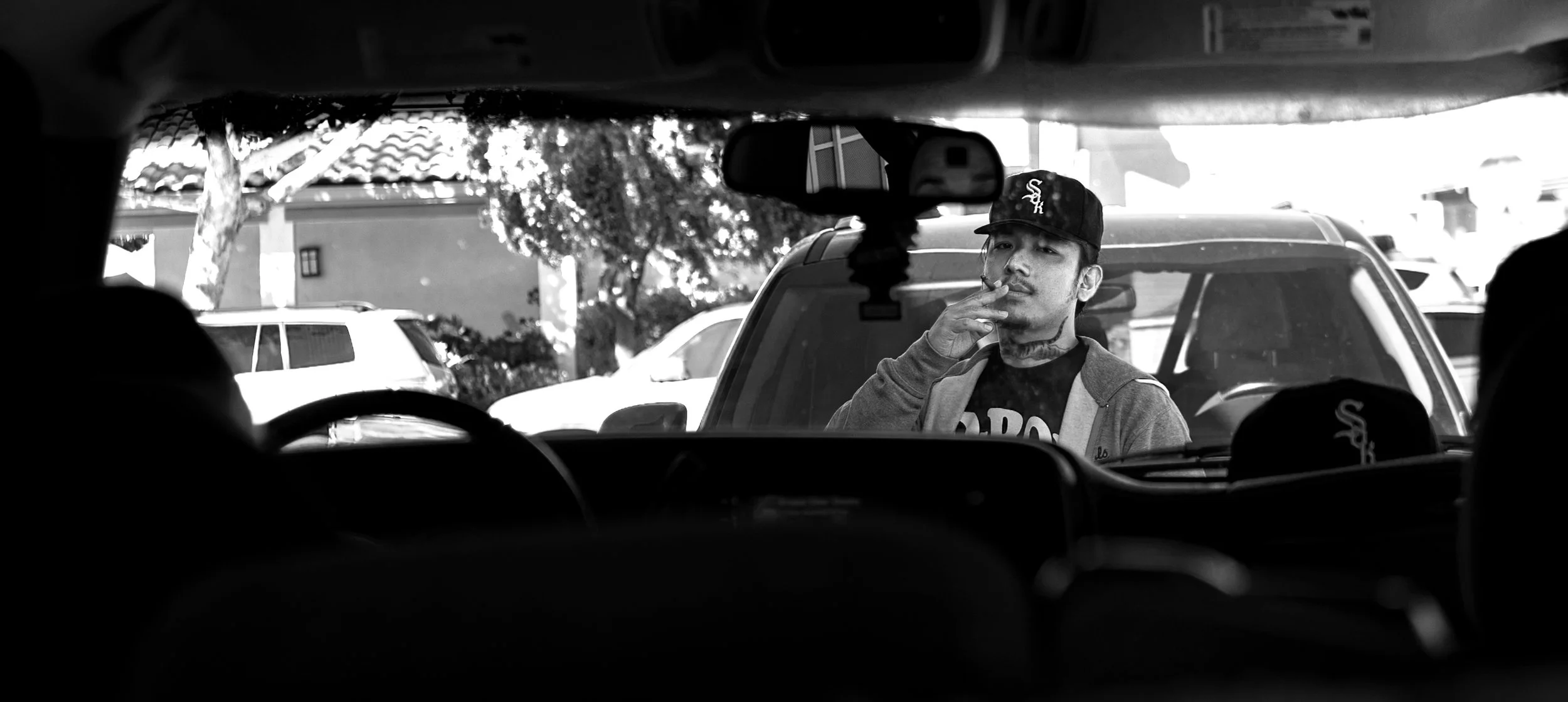 A black and white photo taken from inside a car, showing a man standing outside smoking a thoink. He is wearing a Sok Studios baseball cap and a jacket. There are vehicles and trees in the background.
