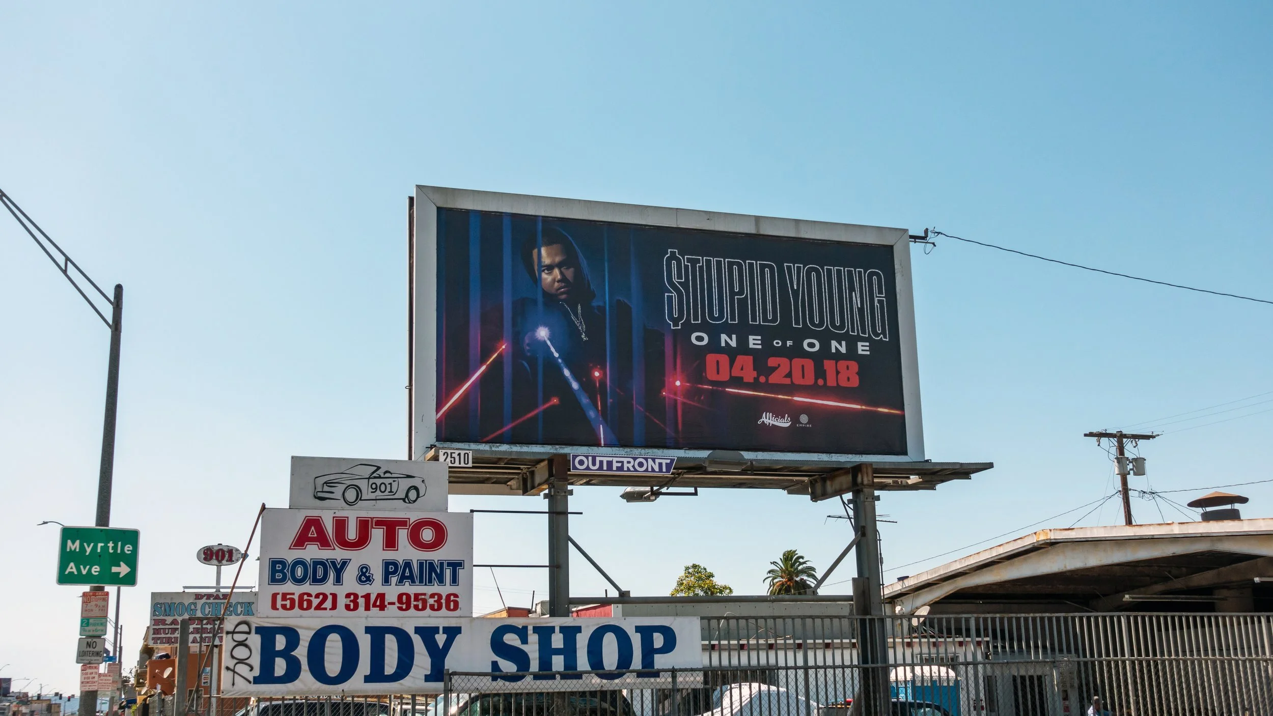 Billboard with advertisement for '$tupid Young - One of One' album release on April 20, 2018, located above an auto body shop on a street corner with visible signage and power lines.