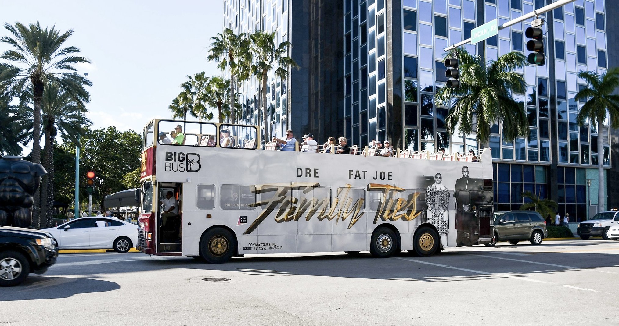 Open-top double-decker bus with "Family Ties" advertisement and passengers on board, in city street with palm trees and modern buildings.