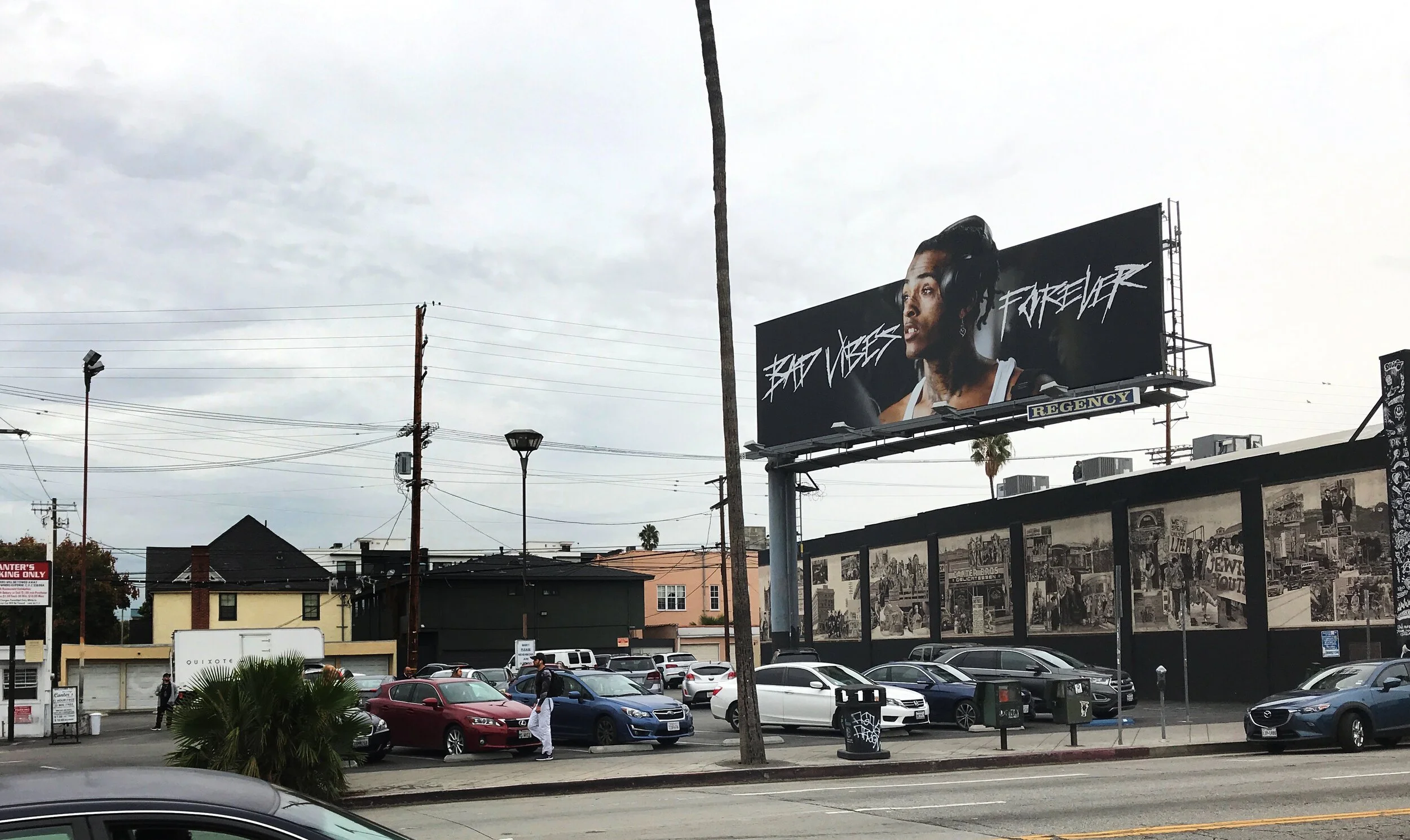 Outdoor parking lot with various cars, a large billboard displaying "Bad Vibes Forever" and a portrait of a person, streetlight poles, and a mural on a wall showcasing historical images.