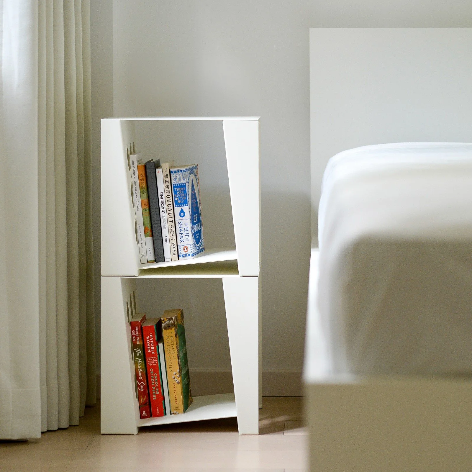 A minimalist white bookshelf with two shelves holding books, placed near a white curtain and a bed with a white sheet in a bedroom.