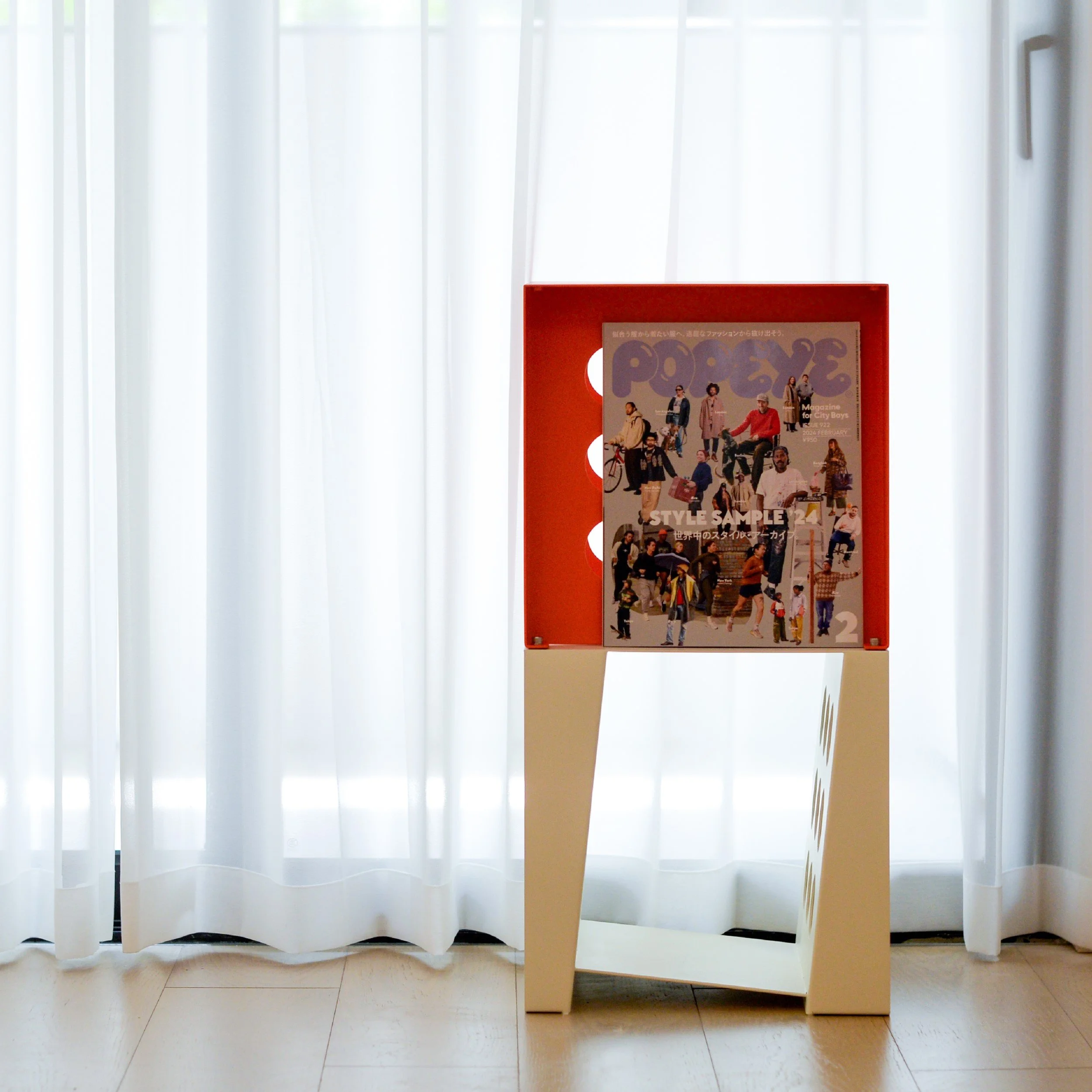 A magazine display stand with a cover featuring various people, set against white curtains with natural light.