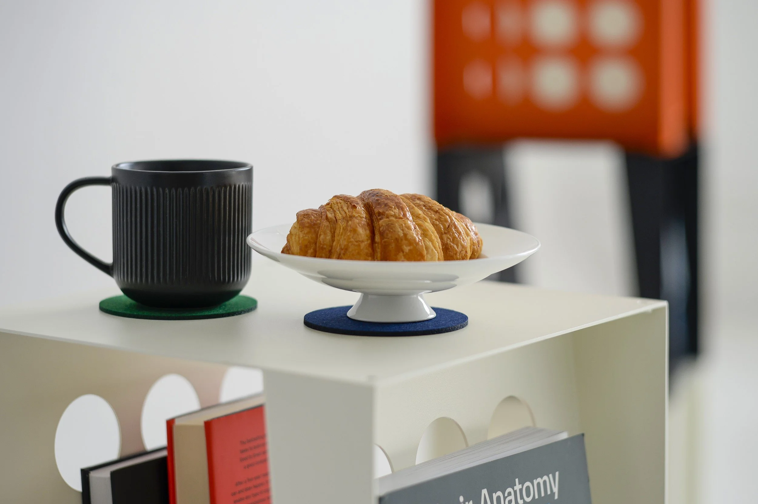 A black ceramic mug on a green coaster, a plate with a croissant on a blue coaster, and a black and orange object in the background, all on a white table.