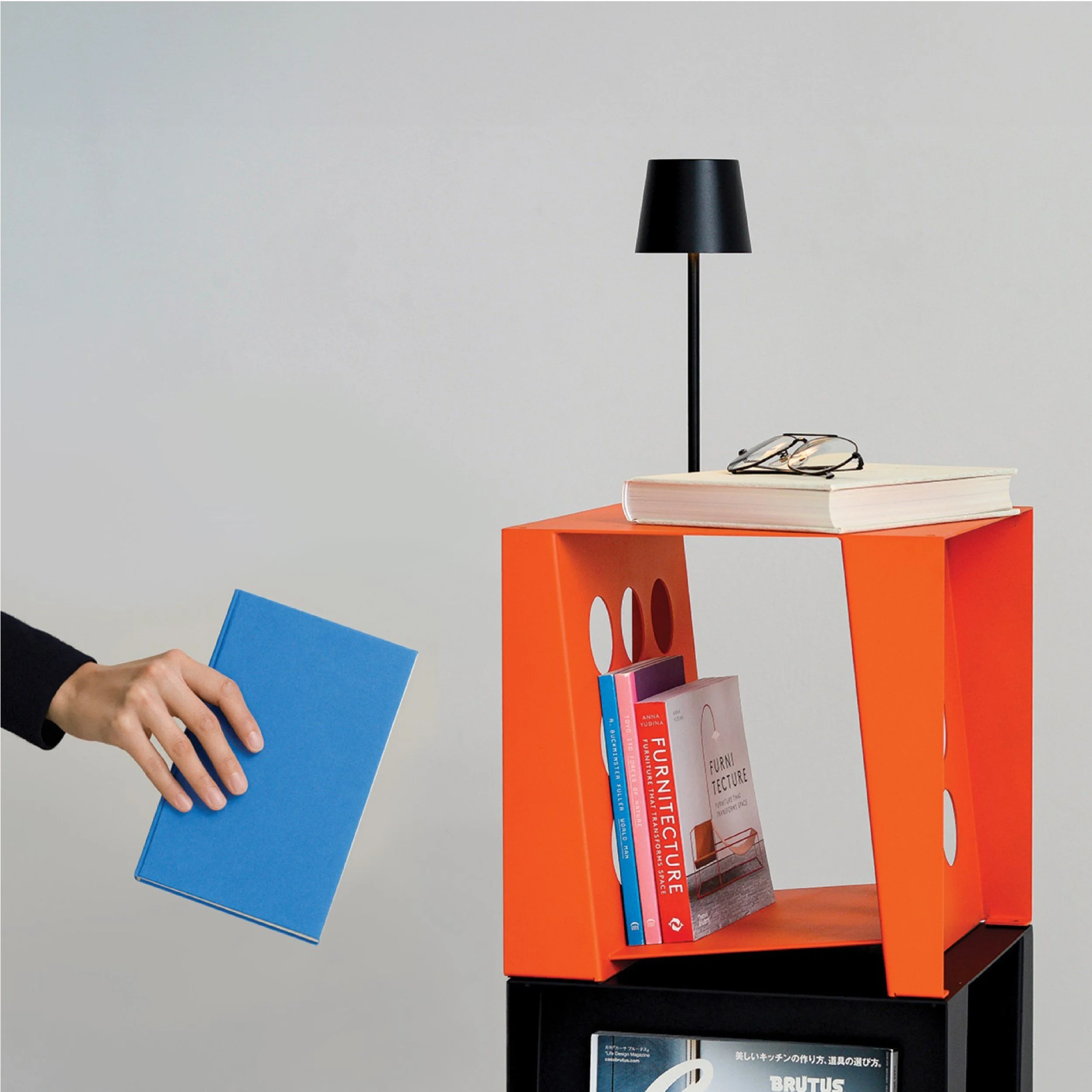 A person holding a blue book in front of a modern orange bookshelf with a black base, decorated with a black table lamp, a white book, glasses, and several other colorful books.