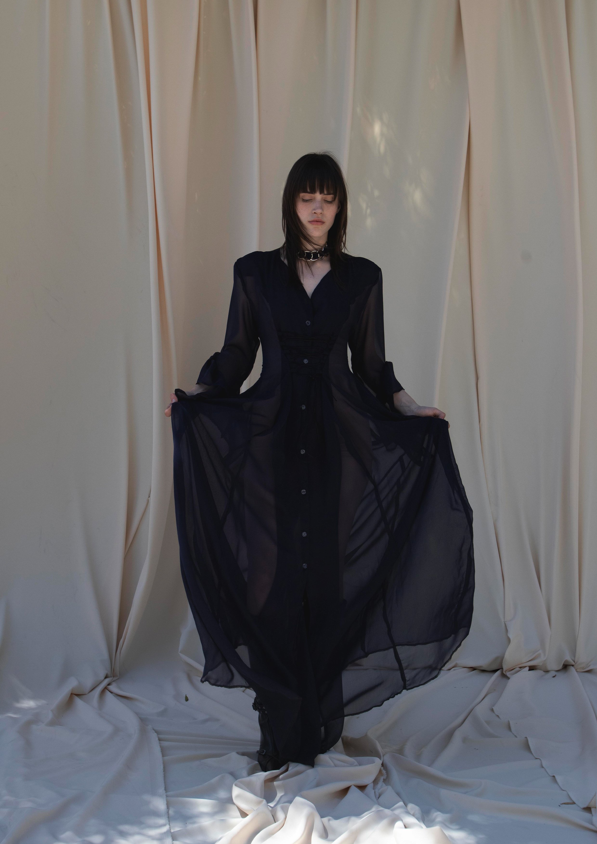 A young woman with dark hair and bangs, dressed in a sheer black gown, standing on a cream-colored fabric backdrop, holding the sides of her dress and looking down with a neutral expression.