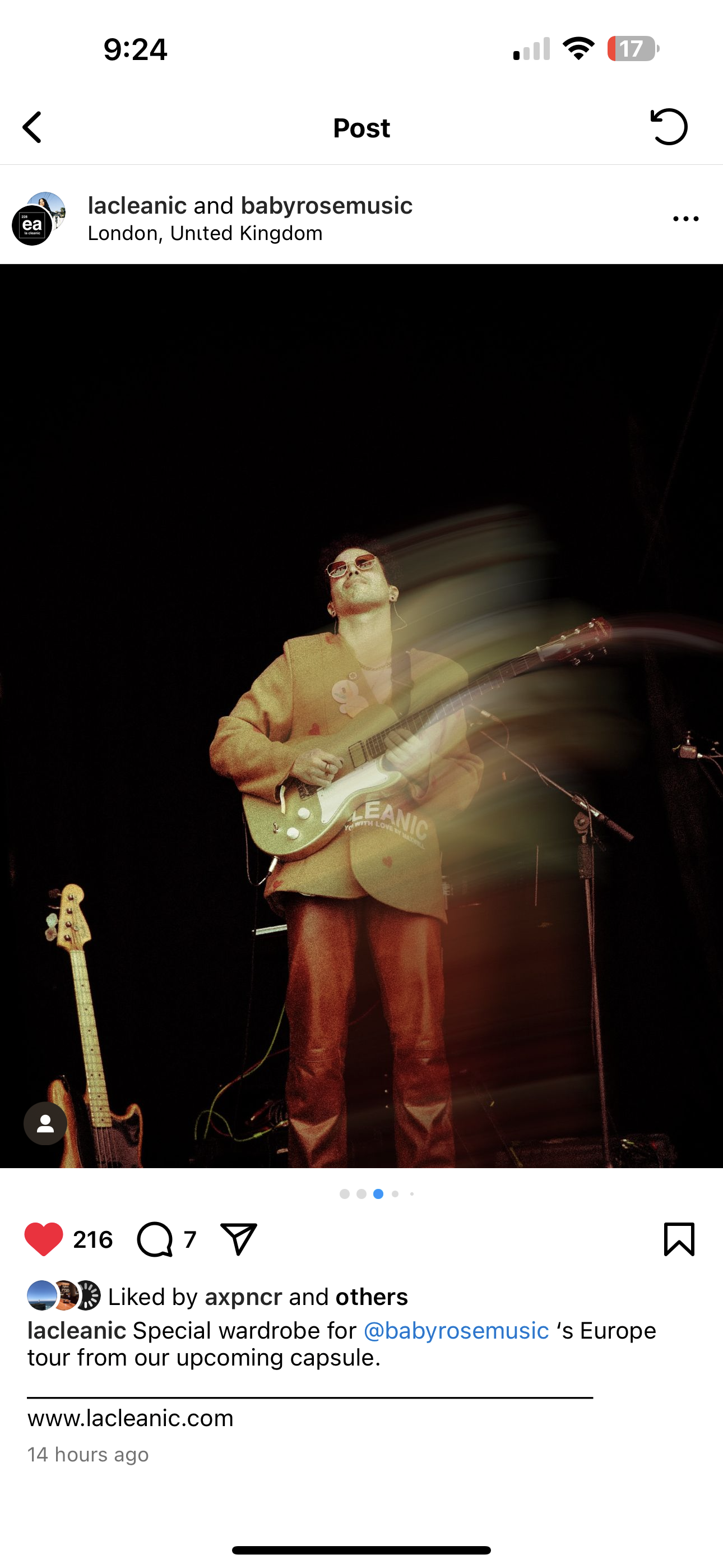 Performer on stage playing guitar with motion blur, wearing sunglasses and a brown blazer, with a second guitar standing on a stand nearby.