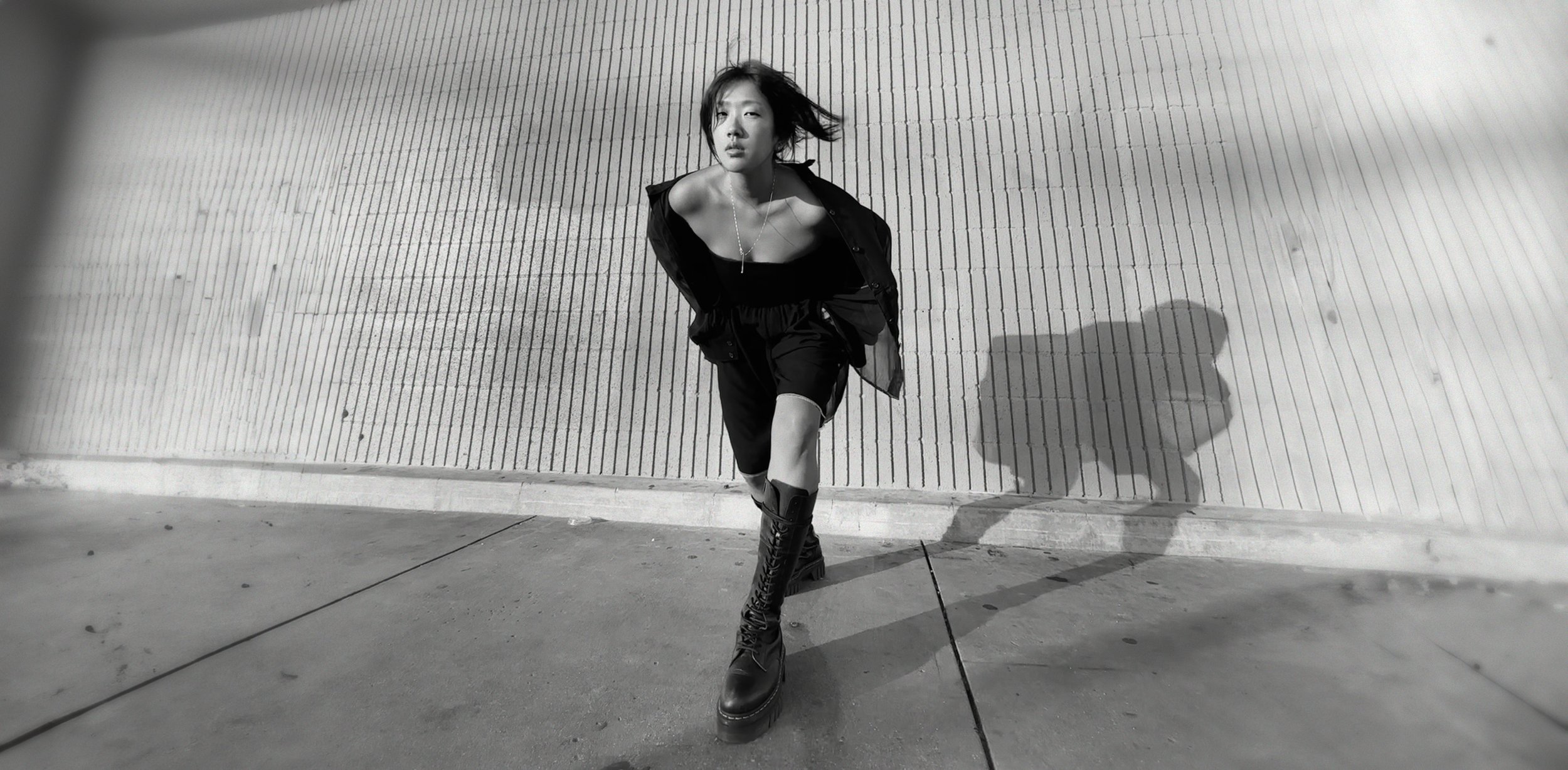 Black and white photo of a woman walking towards the camera against a textured wall, with her hair blowing and casting a shadow on the ground.