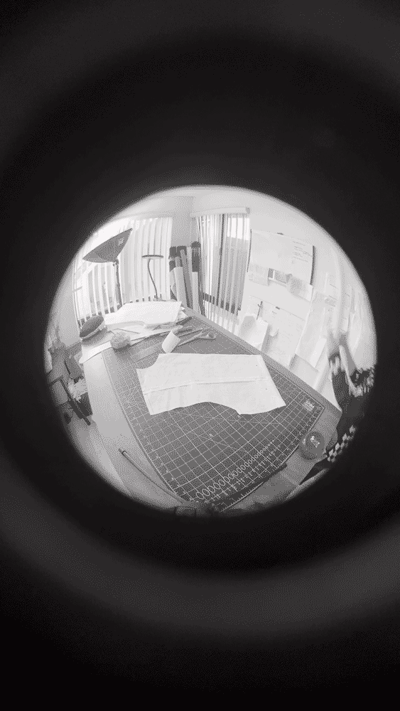 View through a fisheye lens of a workspace with pattern paper, cutting mat, lamp, and some other materials on a desk, with windows and curtains in the background.