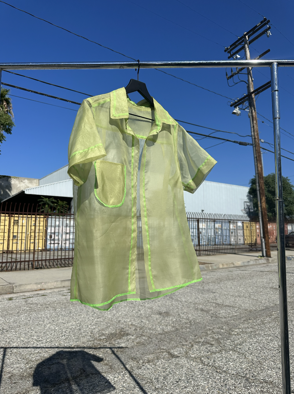 A sheer yellow safety shirt hanging on a black hanger on a metal rod outdoors with utility poles and industrial buildings in the background.